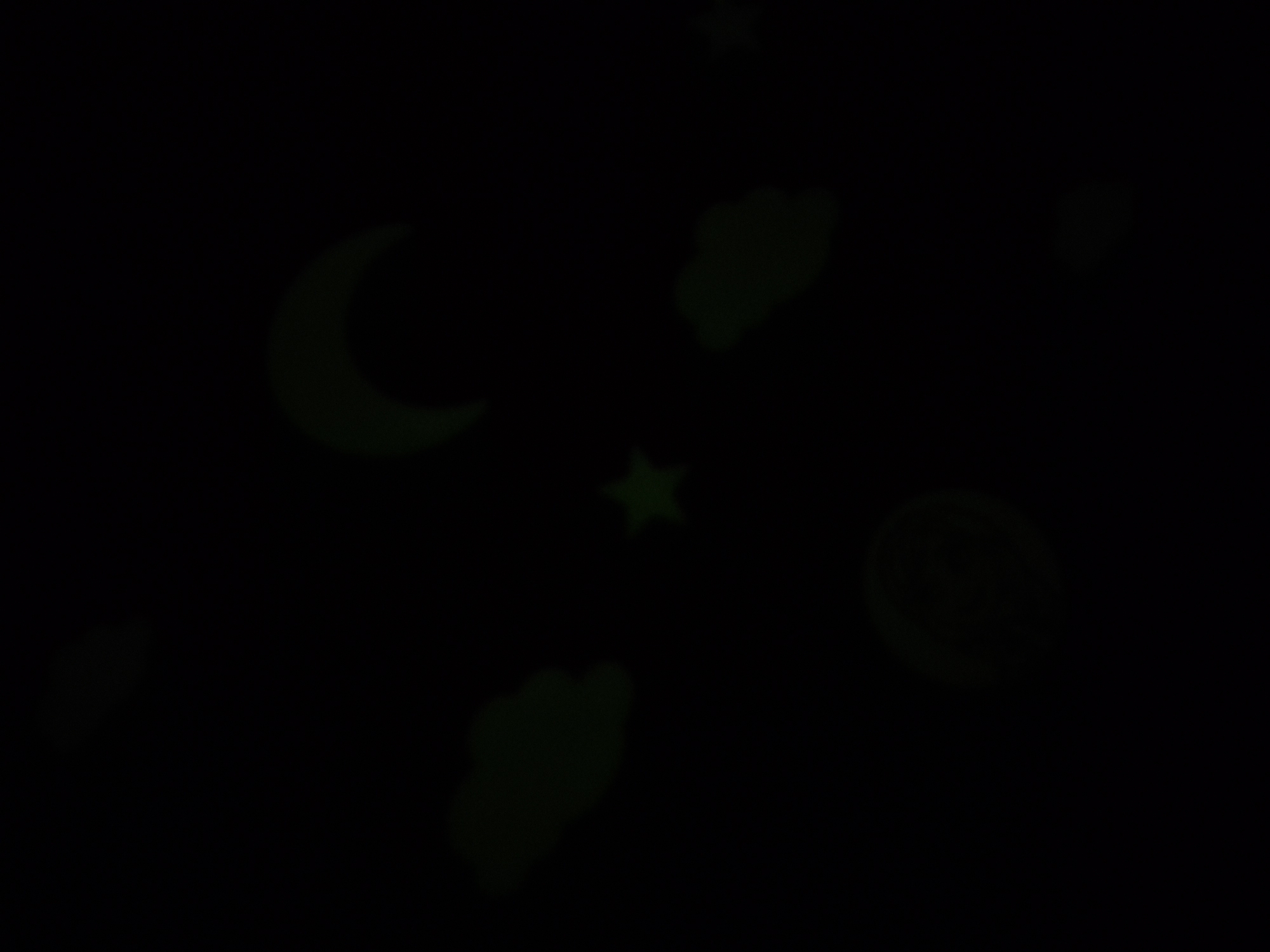 The image displays a dark background with several luminous, greenish shapes scattered across it. These shapes appear to be celestial bodies and clouds, rendered in a stylized, somewhat cartoonish manner. Prominently visible are a crescent moon in the upper right quadrant, a five-pointed star slightly below the center, and several amorphous cloud-like shapes distributed around the frame. There is also a circular object with swirling patterns in the lower center, which might represent a planet or a stylized moon. The lighting is dim, with the shapes emitting a soft glow that illuminates their forms against the dark backdrop. The scene is static, with no discernible action or interaction. The overall impression is of a nighttime sky adorned with glow-in-the-dark decals. No information about the location or context within a city is available.