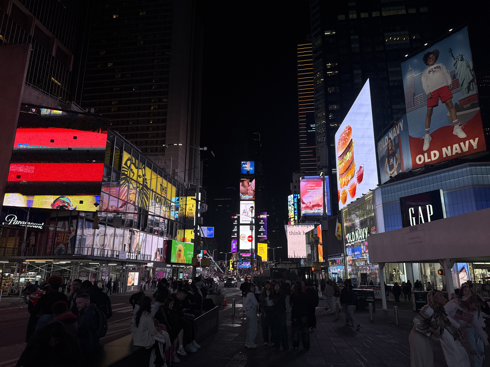 This photograph captures a nighttime scene of Times Square in New York City, United States of America. The foreground is filled with pedestrians, some standing and others seated on benches or curbs. The background is dominated by brightly lit billboards advertising various brands like Old Navy, Gap, and Paramount. These displays are mounted on the surrounding buildings, creating a vibrant and bustling atmosphere. Some of the other brands that can be identified on the billboards are CBS News, Saint, Britbox, Samsung and Arise. The sky is dark and unadorned, allowing the artificial lights of Times Square to stand out. The photo is taken from street level, providing a perspective of the scene as it unfolds to a pedestrian in Times Square.