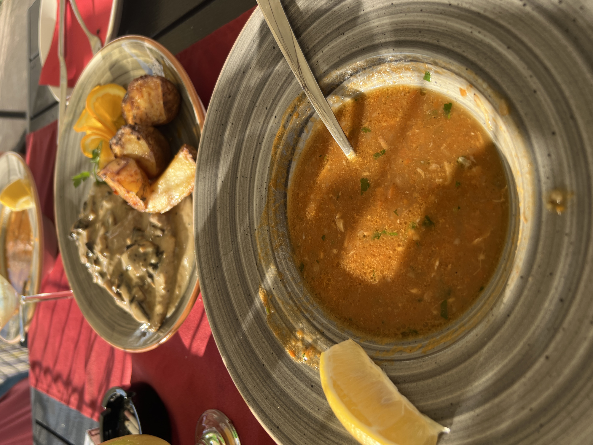 A close-up shot shows a bowl of soup with a lemon wedge on the side, on a grey, textured plate. The soup is a vibrant orange color and appears to have small pieces of meat and vegetables in it. A spoon rests in the bowl. In the background, another plate holds a dish with meat in a creamy sauce, roasted potato wedges, and orange slices. The setting appears to be an outdoor restaurant table with red tablecloths. The image was taken in Sveti Stefan, Montenegro, where the food is served in an outdoor dining area.