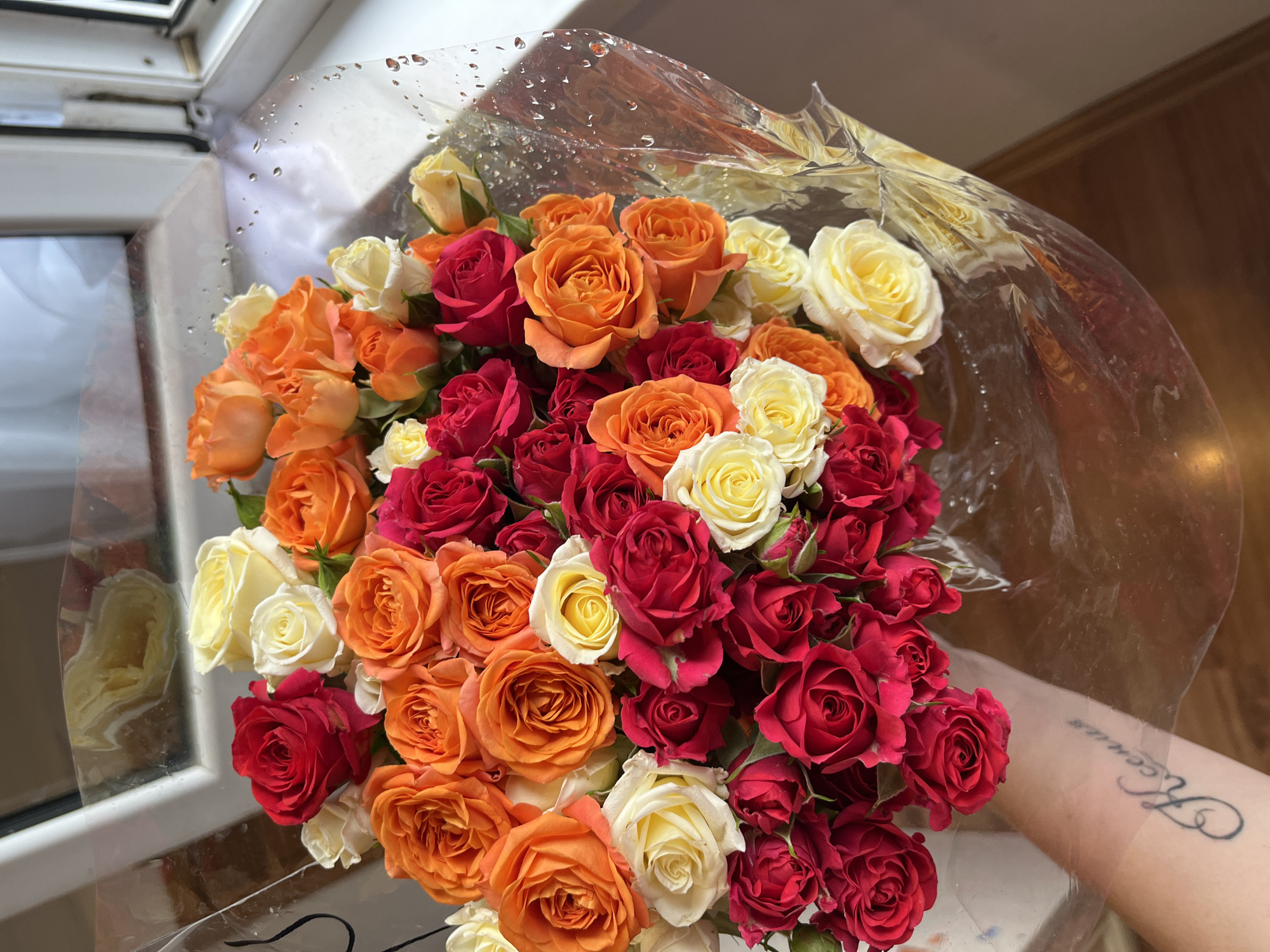 A bouquet of roses in shades of red, orange, and yellow is held by a person's arm with a tattoo. The roses are wrapped in clear plastic, which is slightly damp with water droplets. The bouquet is positioned in front of a window with a view of what appears to be a windowsill and a white frame. The background is out of focus but suggests an indoor setting. The arm with the tattoo is visible in the lower left portion of the image, with the tattoo appearing to be script. The lighting suggests it is daytime. The location is not determinable from the image.