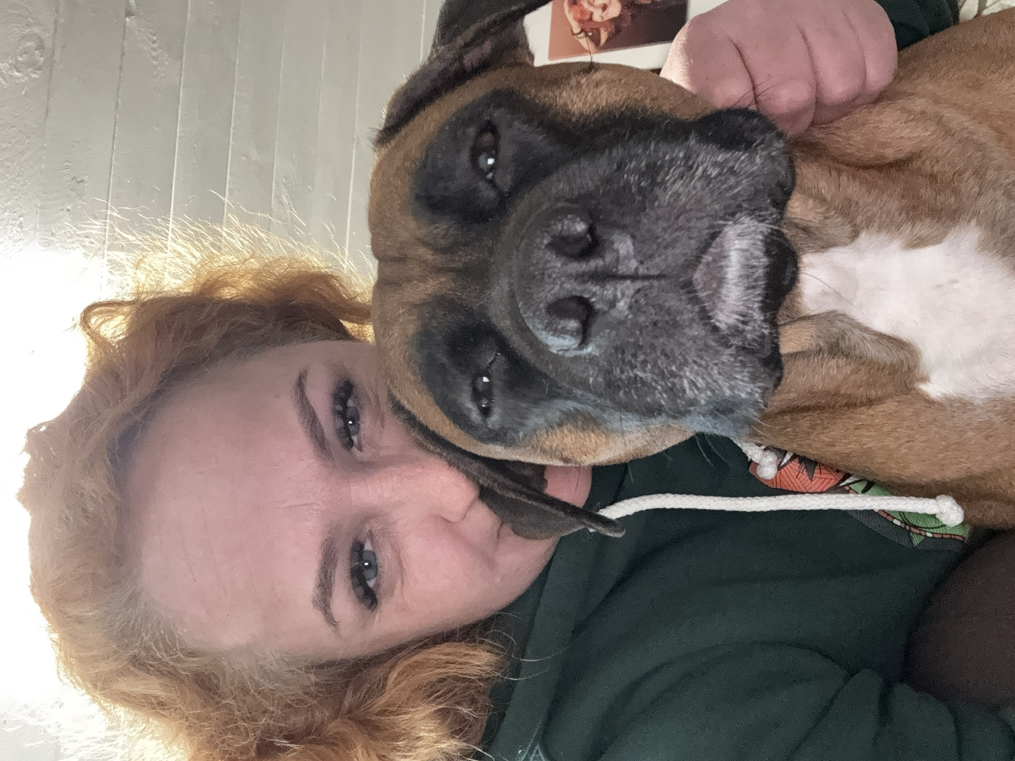 A woman with curly red hair is lying on her back. A brown Boxer dog with a black mask is nestled close to her face. The woman is wearing a dark green hoodie with a white drawstring. Part of a framed image is visible in the background. The location is unknown.