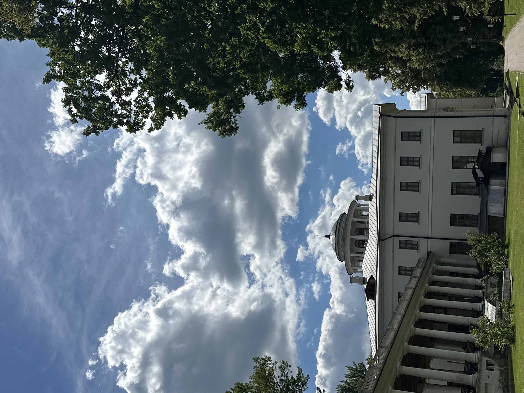 The image depicts the Ostafievo State Museum-Estate in Ostafievo, Russia. A large, white, neoclassical building with a colonnaded portico is the central subject. The building features a prominent dome with arched windows and a spire on top. The sky is filled with dramatic cumulus clouds against a bright blue background, with a large green tree on the right framing the scene. A paved walkway leads into the distance beside the building. The grounds are covered in green grass, with some trees and bushes visible. The image is a full shot with the camera angle slightly tilted upwards.