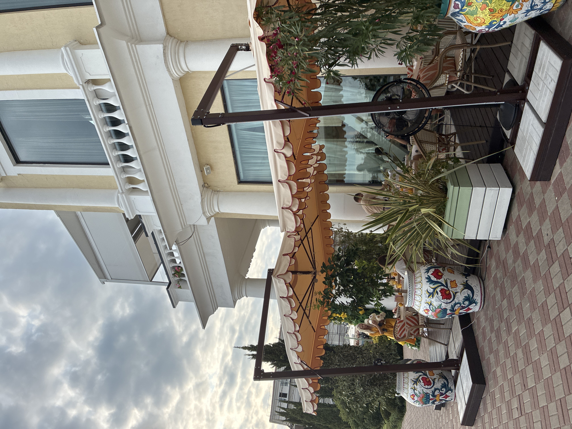 The image depicts an outdoor seating area of a restaurant or cafe in Sochi, Russia, with a large, multi-story building with yellow walls and white columns in the background. Several umbrellas with scalloped edges provide shade over tables and chairs, where a few patrons are seated. Two large, decorative ceramic vases with colorful floral patterns are prominently placed on the paved walkway in the foreground. A woman in a yellow dress is standing near one of the tables, holding stuffed animals. The sky is overcast with scattered clouds. The paving stones are a mix of red and gray.
