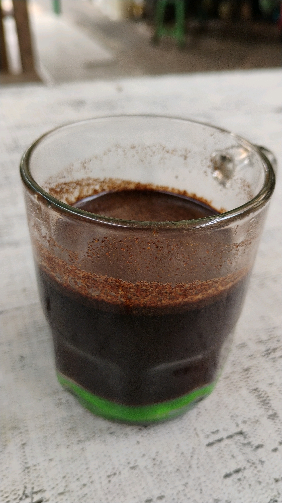A close-up shot shows a clear glass mug filled with dark brown coffee, resting on a white surface. A thin layer of coffee grounds is visible at the top of the liquid, and a subtle green hue is present at the base of the mug. The background is blurred, suggesting an outdoor setting, possibly a cafe or roadside stall in Central Jakarta, Indonesia. The lighting appears to be natural daylight.