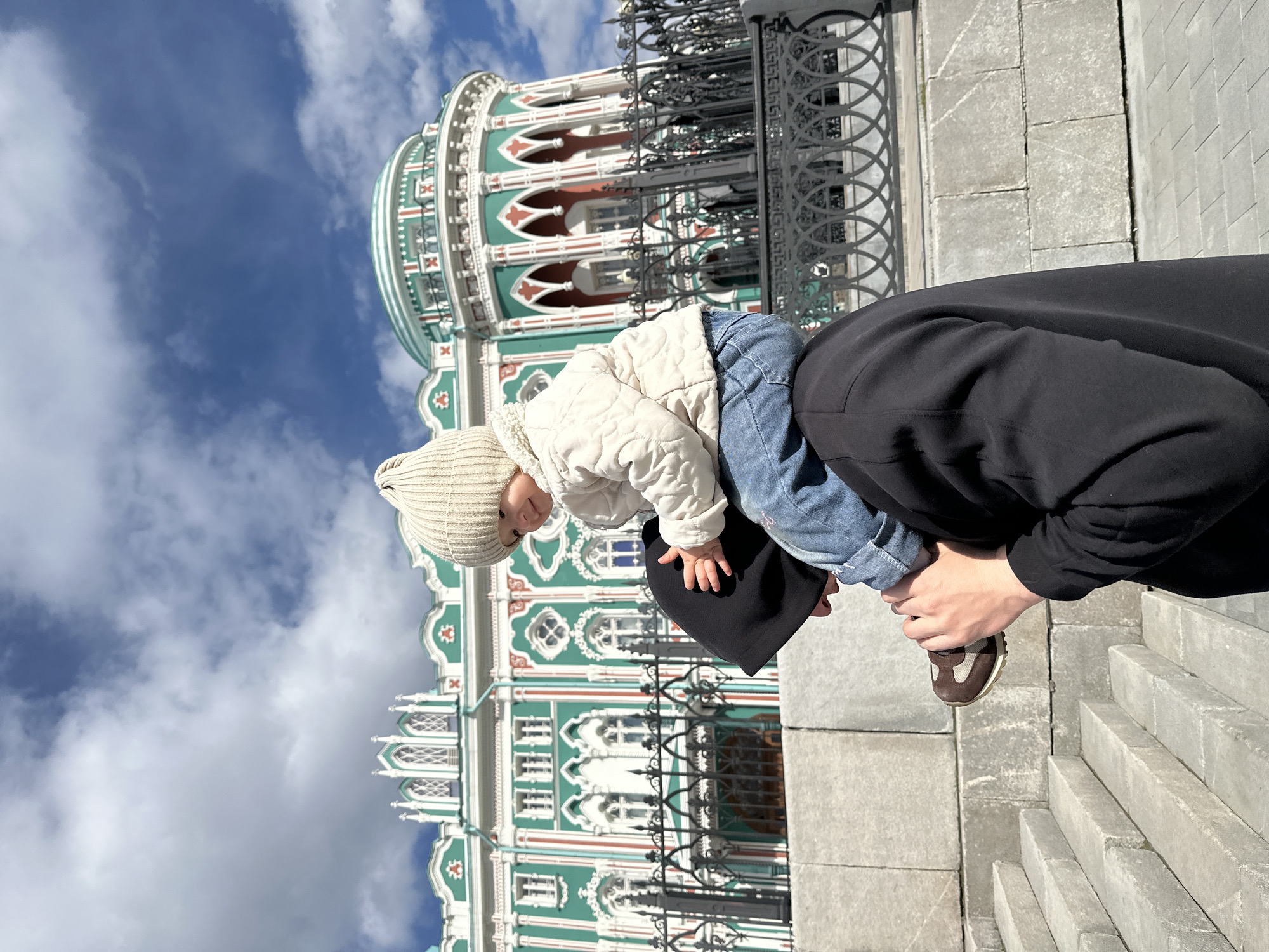 A father is carrying a toddler on his shoulders in Yekaterinburg, Russia. The father is wearing a black hoodie and brown shoes. The toddler is wearing a knit hat, cream-colored jacket, and jeans. In the background is the Sevastyanov House, with its distinctive green and white facade and black ironwork. The sky is visible with blue and white clouds. The father is standing on stone steps.