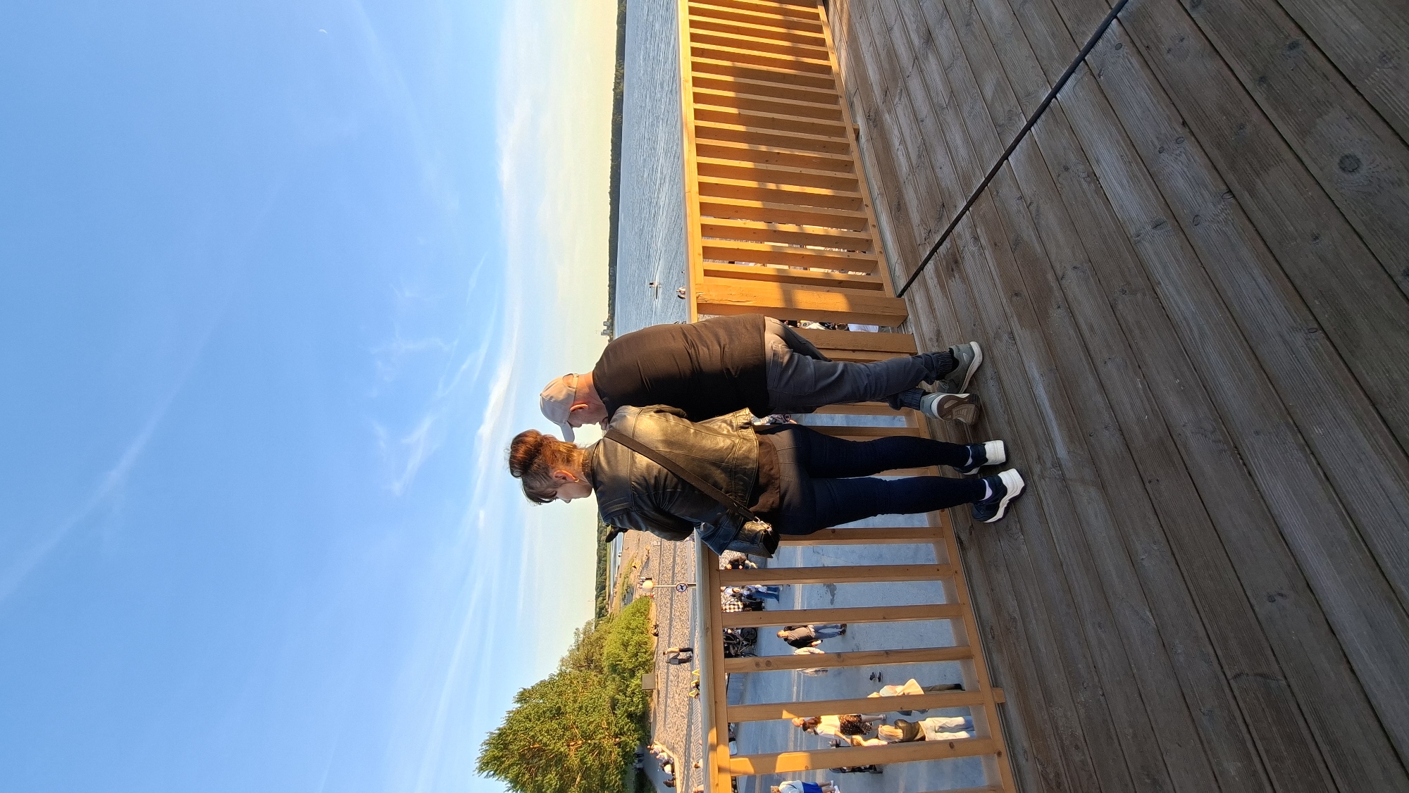 Two people, a woman and a man, stand on a wooden deck, looking out at a body of water. The woman, on the left, wears a black leather jacket, dark pants, and white sneakers. Her hair is pulled up in a bun. The man, on the right, wears a black t-shirt, grey pants, and sneakers, and a light-colored baseball cap. They are positioned side-by-side, with the woman slightly in front.
The deck appears to be part of a waterfront promenade or pier, with a wooden railing separating it from a sandy beach below. On the beach, several other people are visible, some walking and some sitting. Further in the distance, a body of water stretches to the horizon, with a faint treeline on the far side. The sky is a clear blue with some wispy clouds, and the sun appears to be setting, casting a warm glow on the scene.
The location is the Lennusadam Seaplane Harbour area in Tallinn, Estonia, specifically on the viewing platform overlooking the harbor and the beach. The time appears to be late afternoon or early evening.