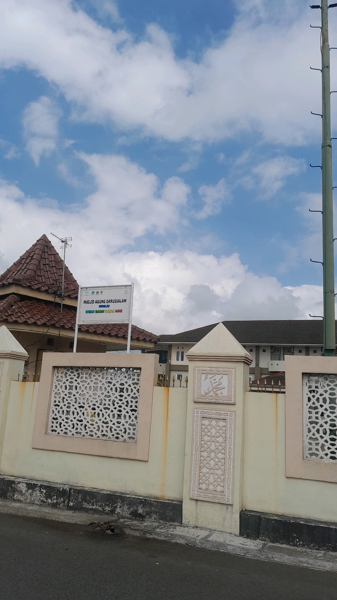 A medium shot captures the front exterior of a mosque complex under a bright, cloudy sky. The building, labeled "MASJID AGUNG DARUSSALAM MIDAJU," is visible behind a decorative wall. The wall features intricate white latticework panels set within a pale yellow stuccoed structure. A vertical pole with hooks stands to the right of the frame, adjacent to the mosque's side. The ground in front of the wall is paved with asphalt, showing signs of wear and tear. The overall scene suggests a public building in an urban or semi-urban environment in Cilacap, Indonesia.