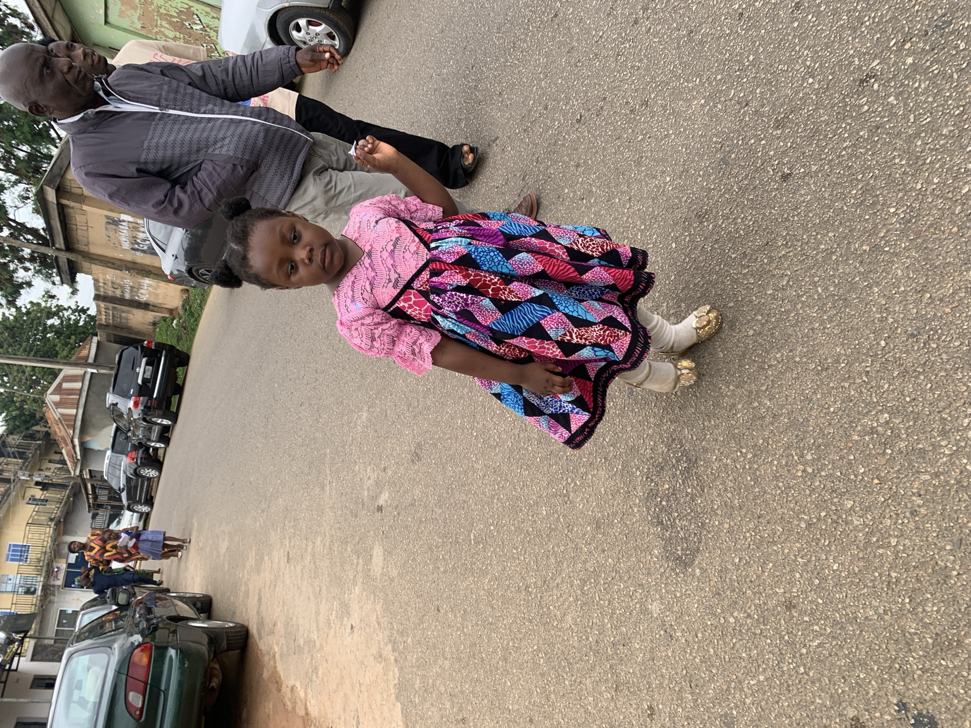 The image captures a street scene in Jos, Nigeria, featuring a young girl in the foreground.
In the center foreground, a dark-skinned girl, estimated to be between three and five years old, stands on an asphalt road. She has dark hair styled into two small buns on top of her head. She wears a short-sleeved dress with a pink lace top and a pleated skirt featuring a vibrant geometric pattern in shades of blue, pink, red, black, and white. Underneath, she wears light-colored leggings and gold-colored flat sandals with a decorative front element. Her right hand is slightly raised, holding a small, folded white object. Her left arm is partially obscured.
An adult dark-skinned male, bald, stands to the girl's right and slightly behind her, appearing to hold her left hand. He wears a grey zippered jacket with a textured horizontal pattern on the chest, a light-colored shirt, grey trousers, and brown shoes. He looks towards the right side of the frame. A younger dark-skinned male, wearing a light-colored long-sleeved shirt, is partially visible behind the first man.
In the mid-ground on the left, another adult dark-skinned woman, dressed in a vibrant orange and yellow patterned outfit, walks along the road. She is accompanied by a dark-clothed man and a second young dark-skinned girl in a light blue skirt and white top, who is holding a piece of paper.
The environment consists of a paved asphalt road, appearing coarse and slightly worn. Vehicles are parked on both sides of the street. On the left, a green sedan (model consistent with a Toyota Corolla) and a grey sedan are visible. On the right, two black SUVs (consistent with Toyota Land Cruisers) and a portion of a silver car are parked. Buildings line the street, including multi-story residential or commercial structures with balconies and a single-story building with a reddish corrugated metal roof on the right. Perimeter walls constructed from concrete blocks, some painted light brown, run along properties. One wall visible in the background has the alphanumeric text "GCM 48" painted in white. Trees and sparse foliage are present in the distant background. The scene is illuminated by natural daylight, suggesting an outdoor setting during the day.