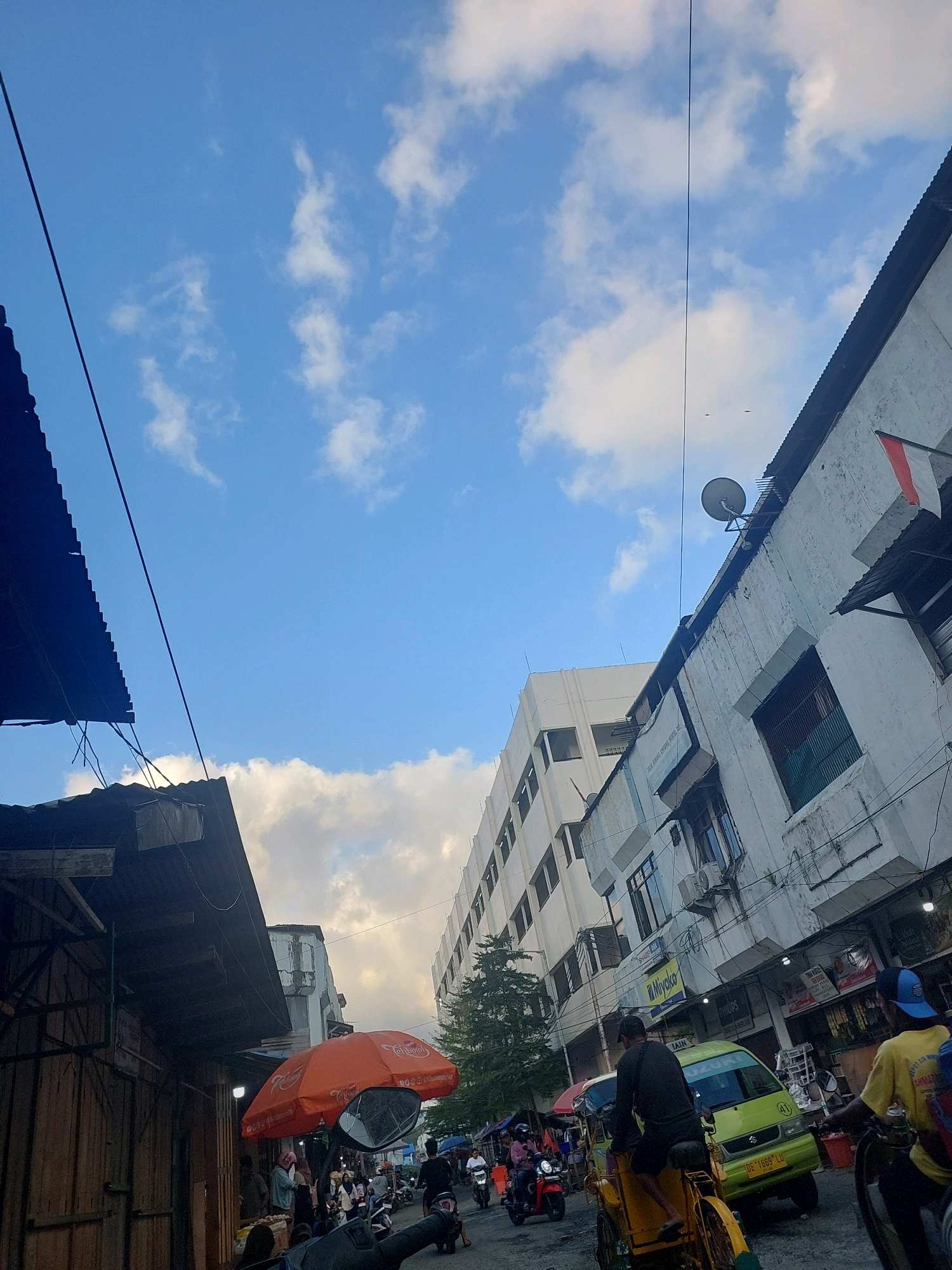 The image depicts a street scene in Ambon, Indonesia, with a focus on the sky and the surrounding urban environment. The sky is a clear blue with scattered white clouds. In the foreground, there is a wooden structure on the left, possibly a market stall, with a corrugated metal roof. A dark blue tarp is also visible. To the right, a multi-story building with a white facade and dark-framed windows dominates the scene. An Indonesian flag, red and white, is attached to the building.
Below the buildings, the street is bustling with activity. Several motorcycles and a small yellow vehicle are present. People are seen walking and riding on motorcycles. An orange umbrella with a logo is open, possibly for shade at a vendor's stall. The overall atmosphere suggests a busy urban setting, likely during daylight hours. The street itself appears to be paved.