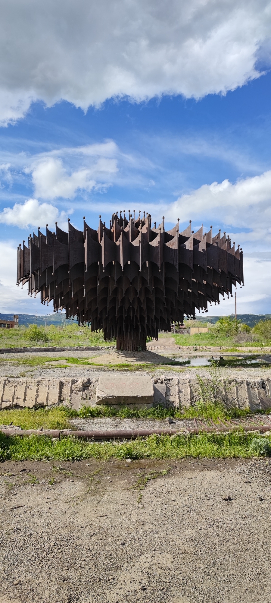 The image captures a desolate scene featuring a decaying metal sculpture in what appears to be a neglected area.
The sculpture, located in Gyumri, Armenia, resembles an abstract tree constructed from multiple layers of dark, oxidized metal plates, each adorned with a small, rounded finial. It stands on a concrete base, surrounded by cracked and weed-infested remnants of a once-maintained area, possibly a fountain or park.
The ground is gravelly and shows signs of disuse. A broken concrete retaining wall borders the area. A rusty pipe lies near the base of the wall. In the background, a few nondescript buildings and green hills are visible. The sky is partly cloudy with patches of blue. The overall atmosphere is one of abandonment and decay. There are no people visible in the scene.