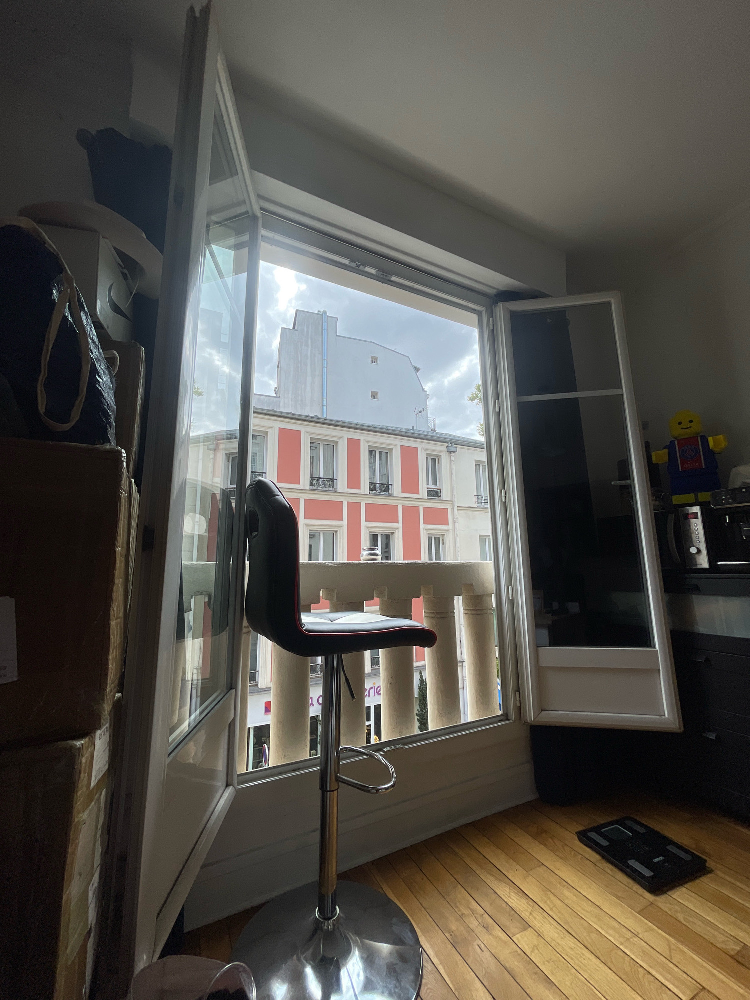 An interior view of a room in Paris, France, looks out through a wide open window onto a street scene. The room has wooden floors and a white ceiling. On the left side of the room, stacked cardboard boxes are visible, along with a black bag on top. In the center of the room, a black and red bar stool stands in front of the open window. The window frame is white, and a portion of a white balcony railing with columns is visible outside. Beyond the railing, apartment buildings with red and white facades, and several windows, line the street. The sky is overcast. On the right side of the room, a black countertop holds a coffee machine and a microwave, with a large Lego figure standing on top of the microwave. A black digital scale rests on the floor near the right edge of the window.