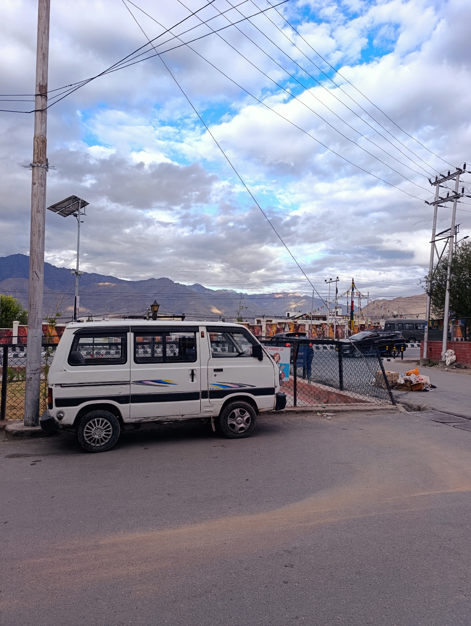 The image shows a white van parked on a paved road in Leh, India. In the background, mountains can be seen, and there are power lines overhead. A tall wooden pole with a solar panel is on the left side of the image, and a chain-link fence is in front of the van. A pile of trash bags is visible next to the road on the right. The sky is partly cloudy.
