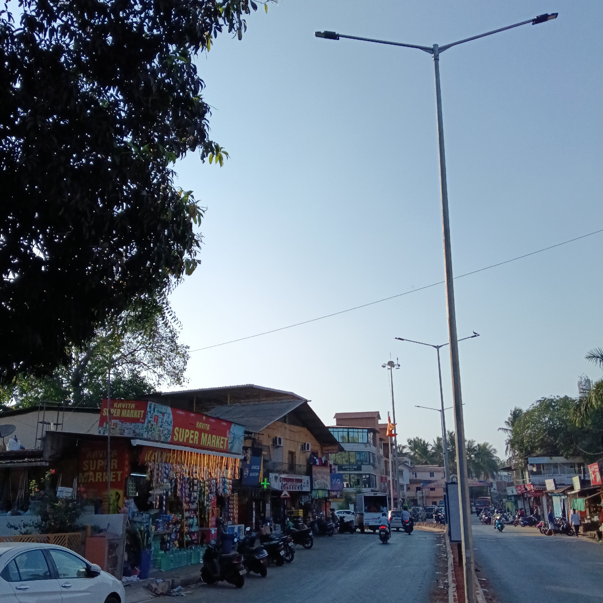 Here is a detailed description of the image:
**General Description:**
The image captures a street scene in Oxel, India, showcasing local commercial activity and urban elements.
**Setting and Environment:**
* **Location:** Oxel, India, as indicated by the local shops.
* **Environment:** A paved street with a row of small businesses on one side. The sky is clear and blue.
**People:**
* There are multiple people visible on the street, mostly riding scooters and motorcycles.
**Objects and Subjects:**
* **Businesses:** Several shops are visible, including:
* "Kavita Super Market" with a distinct red and white sign.
* "Ignelo Bakery."
* "Coiffleur" (presumably a salon).
* "Vivo".
* **Vehicles:**
* Several scooters and motorcycles are parked and moving along the street.
* A white car is parked on the left side.
* A white van is also seen in the middle of the street.
* **Infrastructure:**
* Streetlights are prominent along the roadside.
* Power lines are visible against the sky.
* Buildings with varying architecture line the street.
**Actions and Interactions:**
* Traffic is flowing along the street, with people riding motorbikes.
* Pedestrians are present but not highly visible.
* Shops are open and displaying goods.
**Details:**
* "Kavita Super Market" has a large display of various items outside.
* Several banners and signs advertise the businesses.
**Overall Impression:**
The image depicts a typical street in Oxel, India, bustling with activity, with small shops and local transportation methods being the most prominent features.
