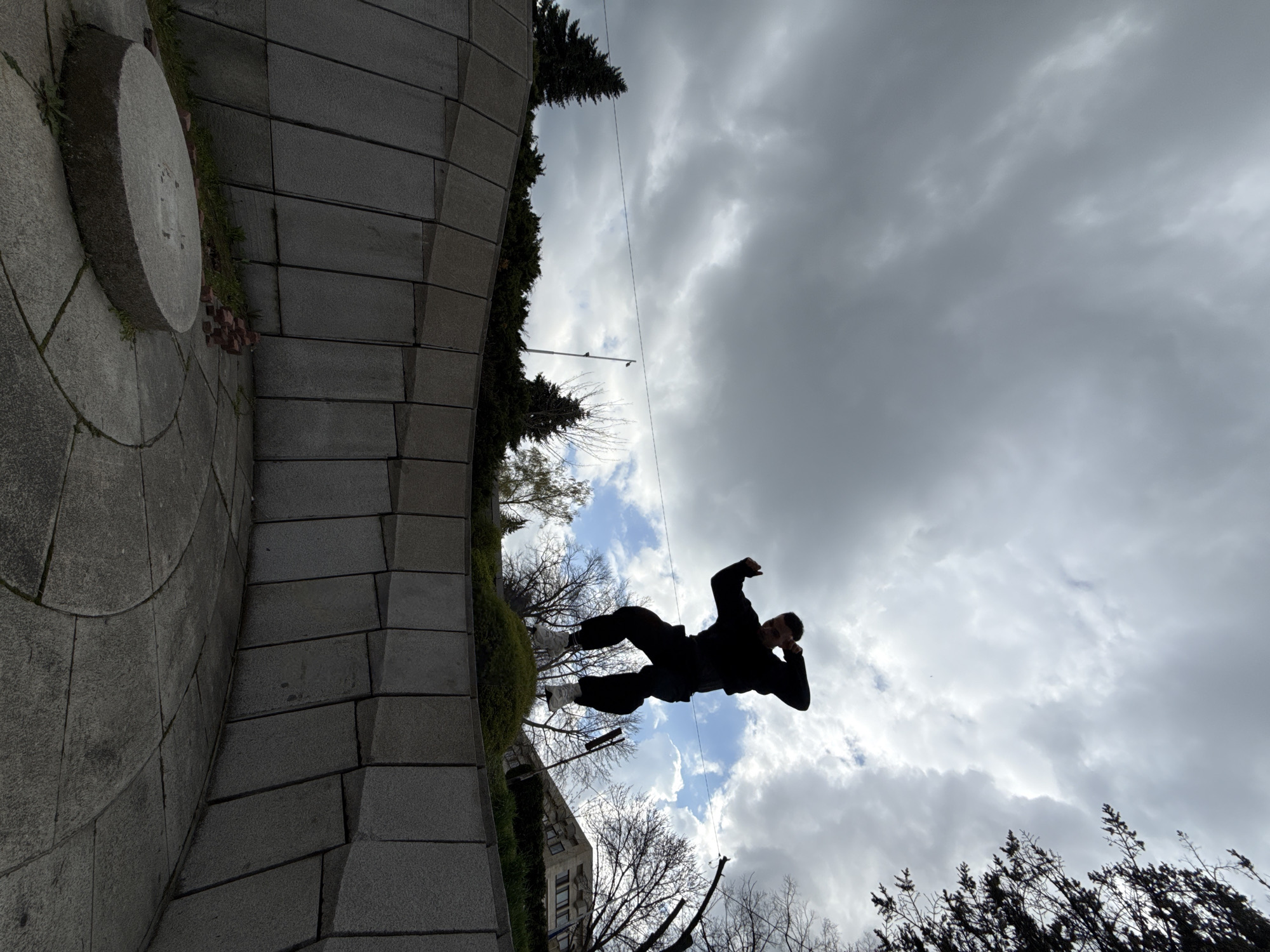 A man is captured mid-jump against a cloudy sky in Shumen, Bulgaria. The image appears to be taken from a low angle, looking upwards. The man is silhouetted, suggesting that he is backlit. He is positioned over a paved structure, which could be part of a monument or public space. Trees and other vegetation are visible in the background and to the side of the paved structure, partially obscuring buildings. The composition also contains electrical wires running across the sky.