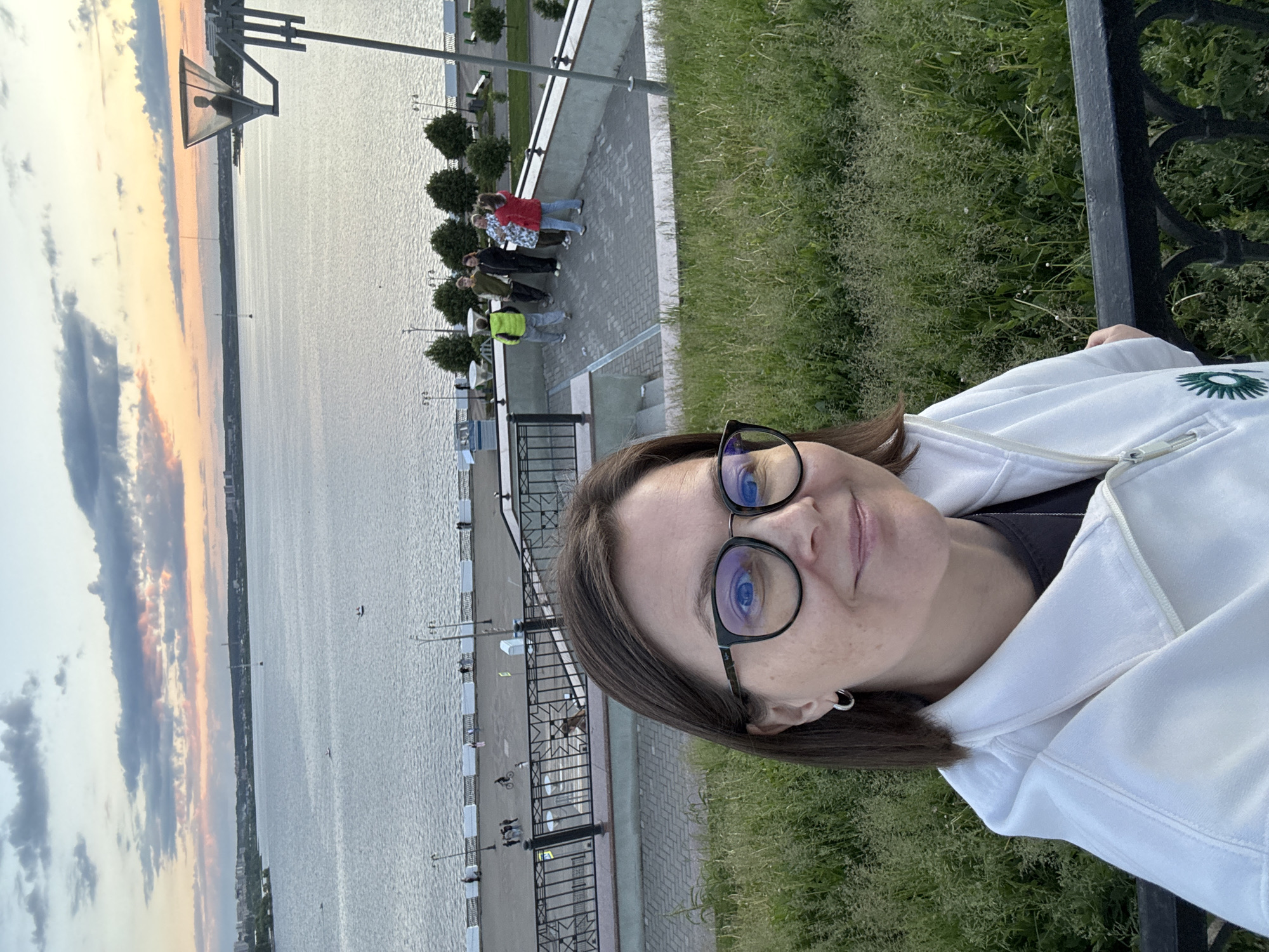 A medium-shot selfie captures a woman with dark hair and glasses, wearing a white hooded jacket, smiling softly. The image is rotated 90 degrees counter-clockwise, giving a horizontal perspective to the main subject. A black metal railing is visible to her right. On her white jacket, a green, stylized sun-like emblem is present, though no clear text is discernible.
The background reveals a scenic waterfront promenade alongside a large body of water, which is the Izhevsk Pond/Reservoir in Izhevsk, Russia. The promenade is paved and lined with streetlights and manicured evergreen shrubs. Several people are visible in the mid-ground, walking or standing on the promenade, including a group of approximately five individuals.
The sky, displaying a blend of orange, pink, grey, and blue hues with clouds, suggests either a sunset or sunrise, with the warm colors indicative of evening. The water reflects the soft light, and distant land or a cityscape can be seen across the expanse. A distinctive lamppost with a square light fixture is prominent on the left side of the promenade. The scene depicts a serene moment at a public recreational area, with calm weather conditions.
