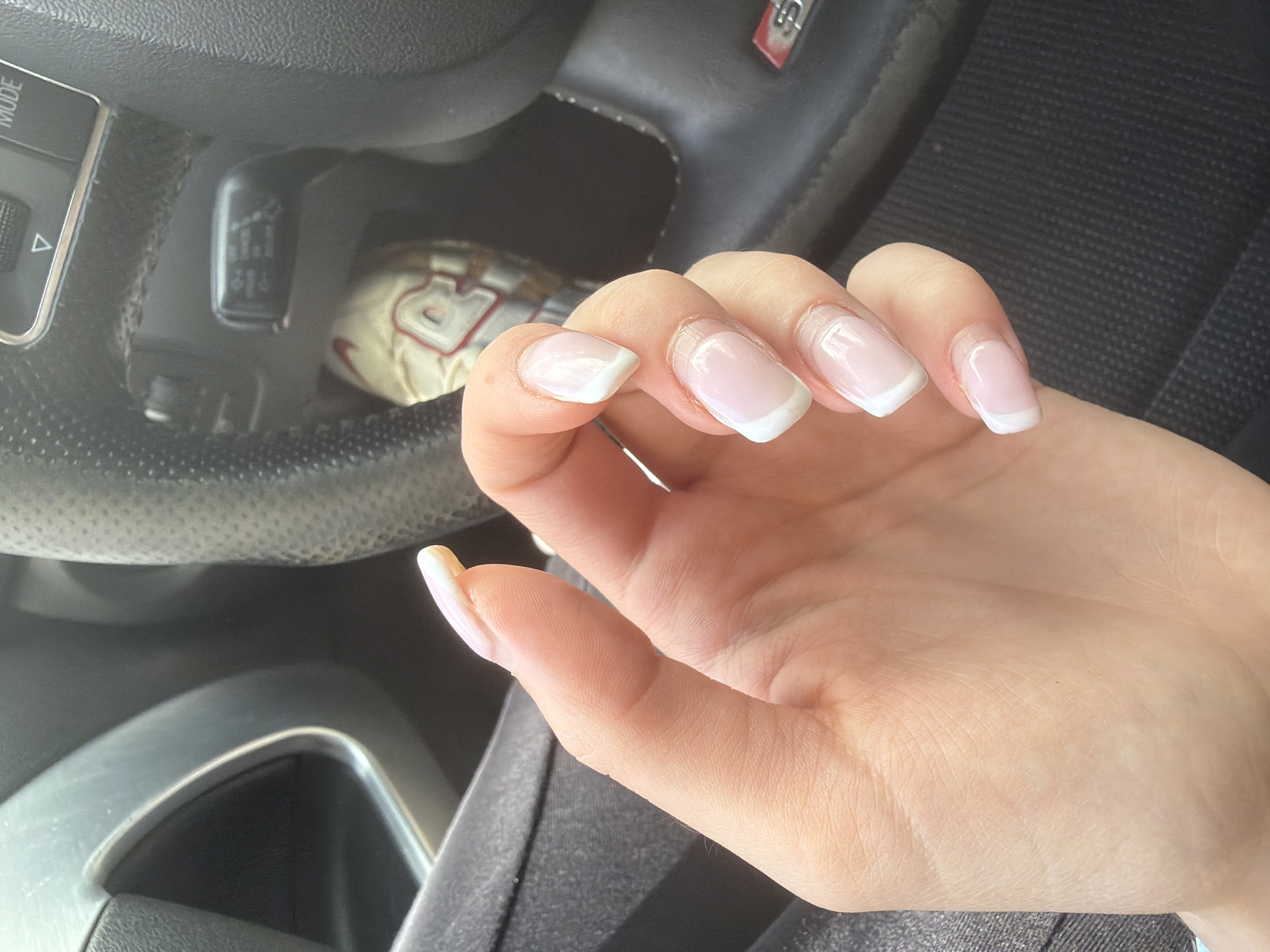 A close-up photograph shows a person's hand with manicured nails exhibiting a white-tipped French manicure. The hand is positioned over a vehicle's steering wheel, which is visible in the background. Part of a car seat is also visible. A partially visible Nike sneaker is in the background. The location is not discernible from the available visual information.