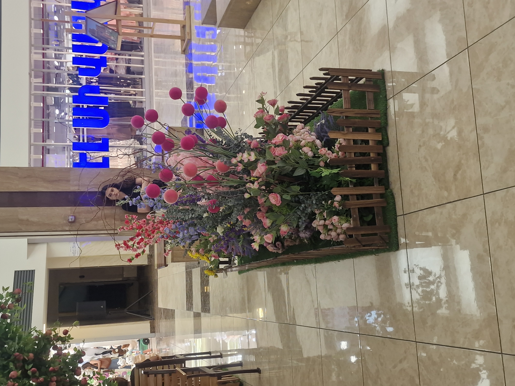 A young woman with dark hair is sitting behind a decorative floral arrangement in what appears to be a shopping mall in Yerevan, Armenia. The floral arrangement consists of various artificial flowers in shades of pink, purple, and red, with some spherical red ornaments. A small wooden fence encloses the base of the arrangement. The woman is wearing a dark t-shirt and is looking to her left. In the background, a store with blue lettering that reads "LU LUX" is visible through a glass facade. The floor is tiled with a beige and brown checkered pattern. To the left of the floral arrangement, a portion of a tree with red fruit is visible. The environment suggests an indoor public space, possibly a mall or a large retail establishment.
