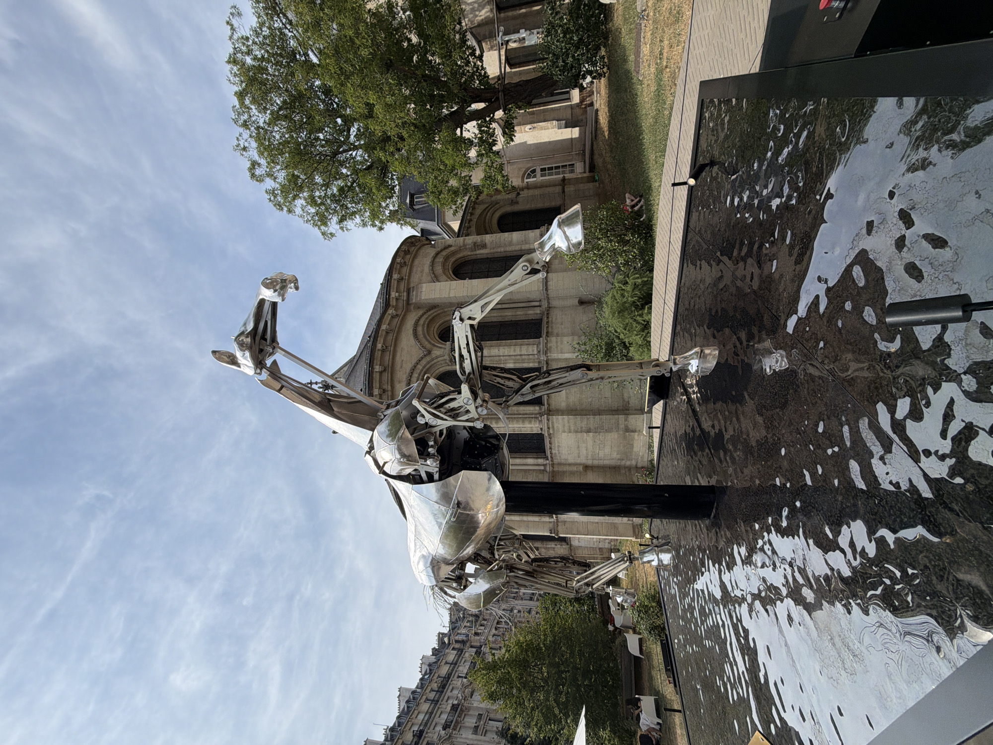 A full shot, eye-level, outdoor photograph captures a large, polished metal sculpture of a horse standing in a shallow pool of dark, rippling water. The sculpture is highly detailed, with exposed internal mechanisms and a skeletal appearance. It is positioned in a courtyard with a light-colored stone building featuring arched windows in the background. To the left of the building, a cluster of trees and a residential building with ornate architecture are visible under a bright, partly cloudy sky. In the foreground, a part of a black base with red and black buttons is visible on the right side of the frame. To the left, people are seated on white lounge chairs in a grassy area, with a white umbrella providing shade. A few more white chairs and a low hedge are also visible in the background. The sculpture's reflective surface mirrors the sky and surrounding environment. The overall setting appears to be a public space in a city, likely a courtyard or garden adjacent to a historical building, potentially in Paris, France.