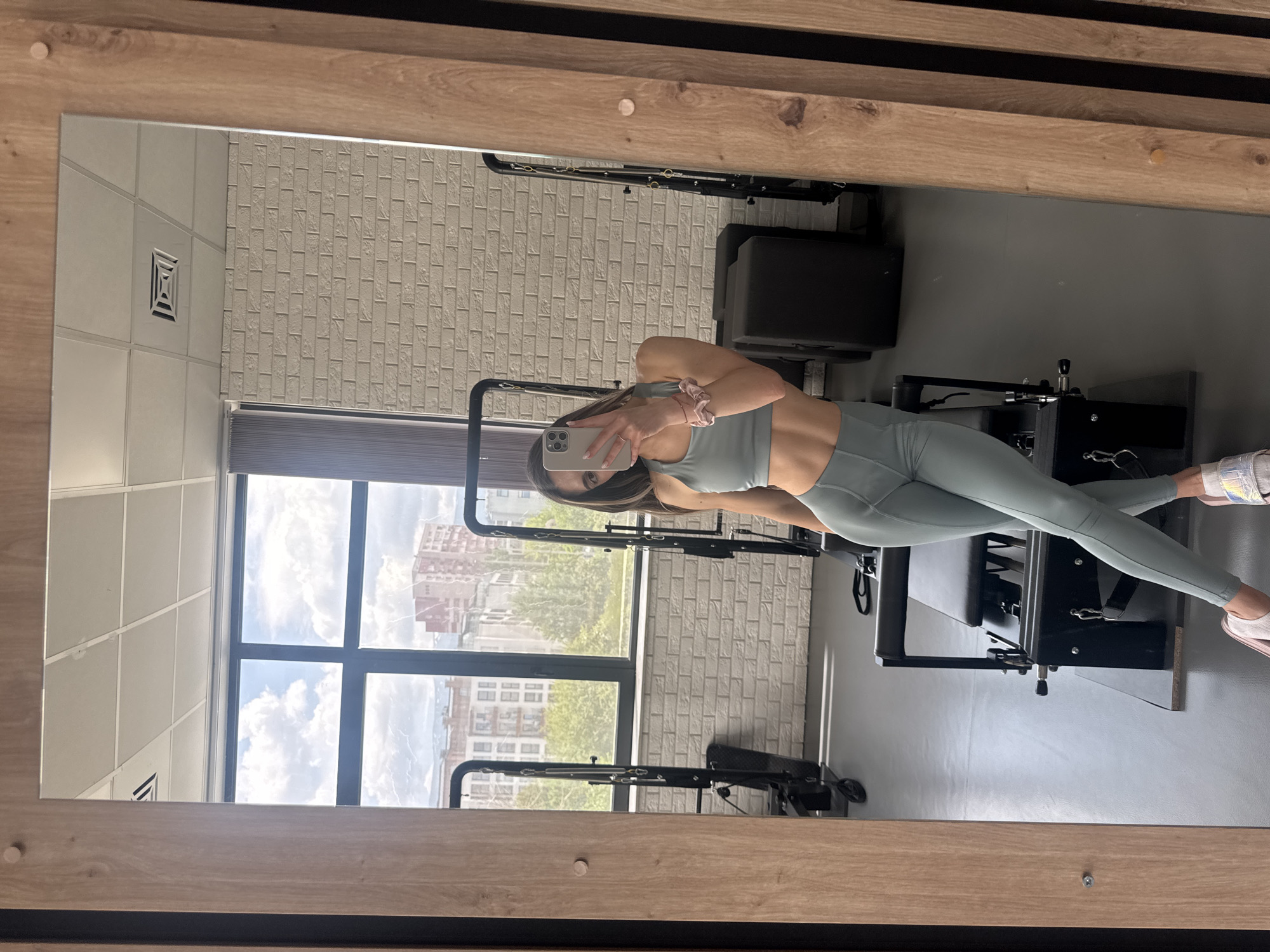 A mirror in a gym setting shows a woman in athletic wear taking a selfie with her smartphone. She is standing on or next to a black pilates reformer machine. The background includes a brick wall, a window with a view of buildings and a blue sky with clouds, and other gym equipment. The location could be Sofia, Bulgaria.