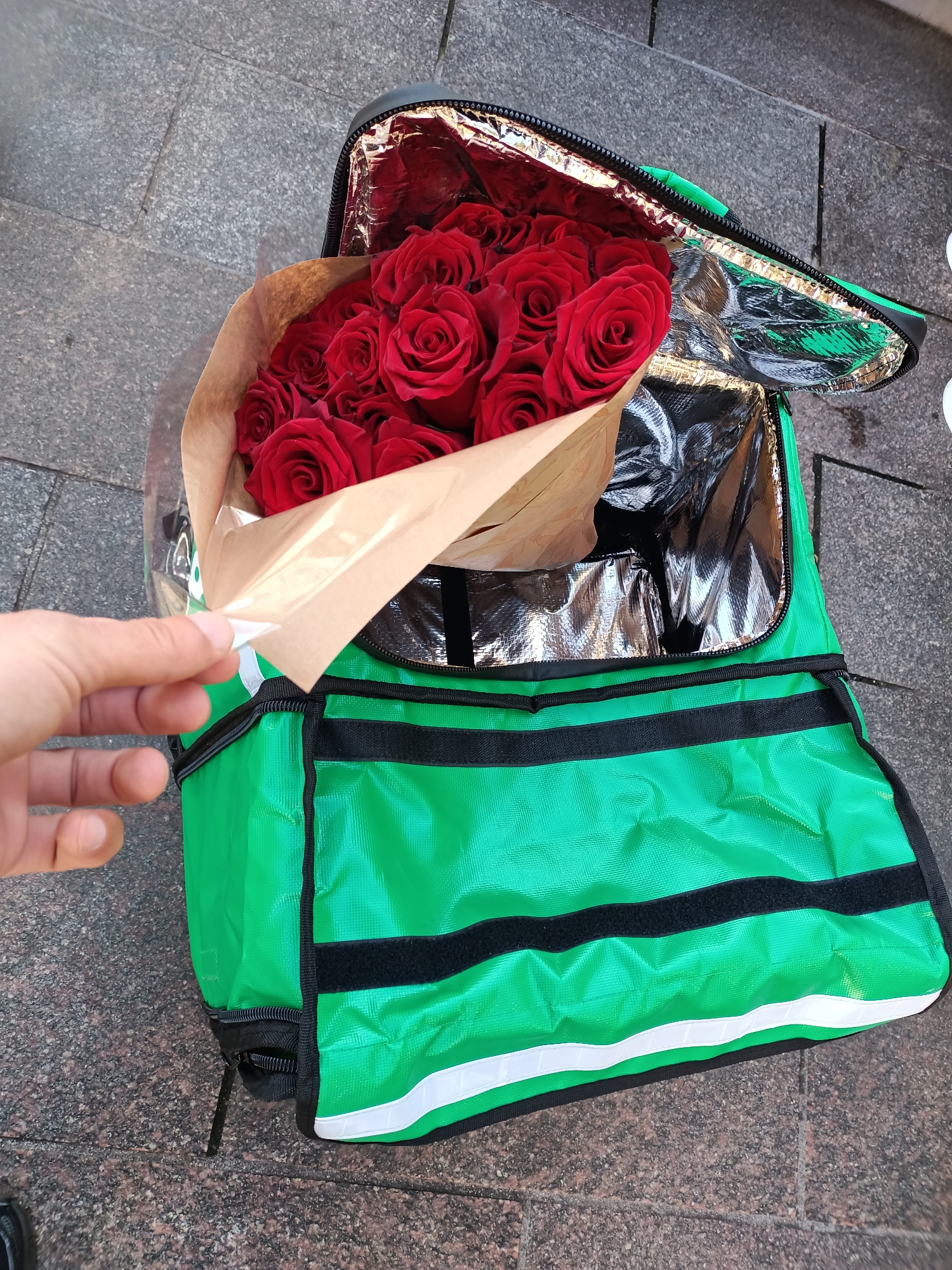 A bouquet of red roses, wrapped in brown paper and clear plastic, is partially inserted into a bright green insulated delivery bag with a black zipper. The bag's interior is lined with reflective material. A person's left hand is visible, gently holding the paper wrapping of the roses near the opening of the bag. The bag rests on a stone-paved surface, likely a sidewalk, with visible seams between the paving stones. The scene appears to be outdoors, possibly in an urban environment. The background is blurred but suggests a city setting. The image is taken from a high angle, looking down into the bag. There are no visible landmarks to pinpoint the exact location within Tallinn, Estonia.