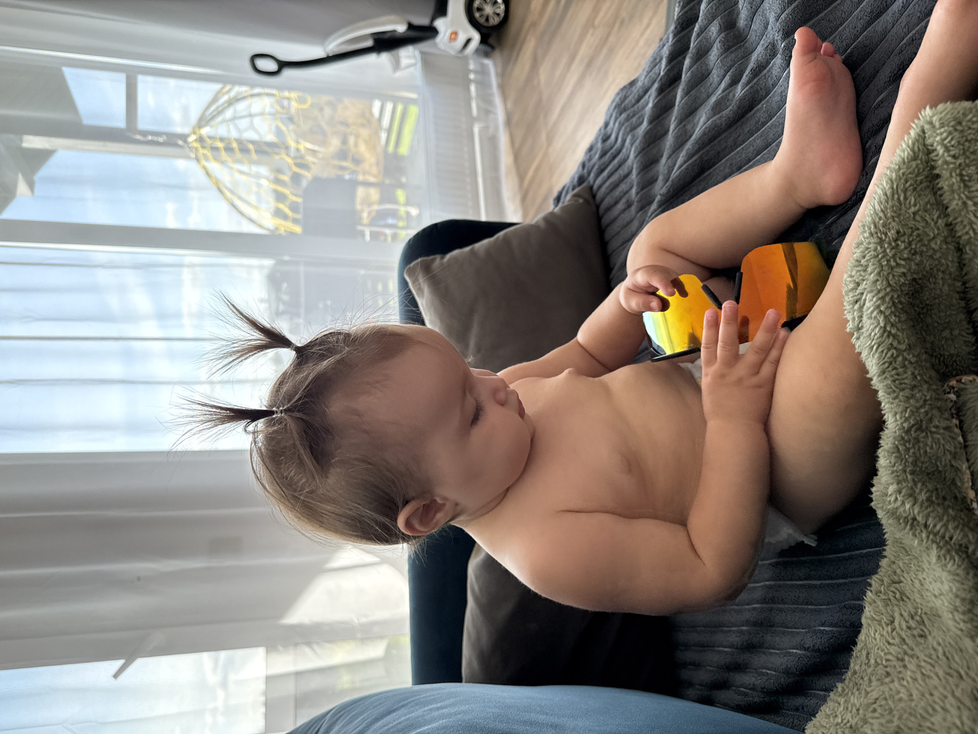 A baby, possibly a toddler, is lying on their side on a striped grey blanket, with a fuzzy green blanket draped over their legs. The baby is mostly undressed, wearing only a diaper. They are holding two brightly colored toy blocks, one orange and one yellow, in their hands and appear to be looking at them with curiosity. The baby's hair is styled into two small pigtails that stick up from their head.
The setting appears to be indoors, near a large window with white blinds. Through the window, a bright, sunny sky is visible, suggesting it is daytime. Outside the window, there are some indistinct structures, possibly part of a building or an outdoor play area, with some yellow framework visible. The floor is wooden, and other furniture, including a dark blue chair and a grey cushion, are visible in the background. A small, white toy vehicle with a wheel is also perched on top of the blinds. The overall impression is of a relaxed, home environment. The baby's expression and posture suggest contentment and engagement with their toys.