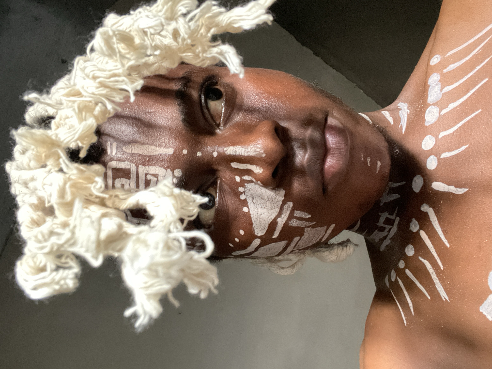 A person with dark skin is facing the camera at an upward angle, with white body paint and wearing a white yarn-like headdress. The body paint consists of geometric patterns and dots on the face, with lines and dots extending down the neck and shoulder. The background appears to be dark and nondescript. No specific landmarks or features indicate a location in Onitsha, Nigeria.