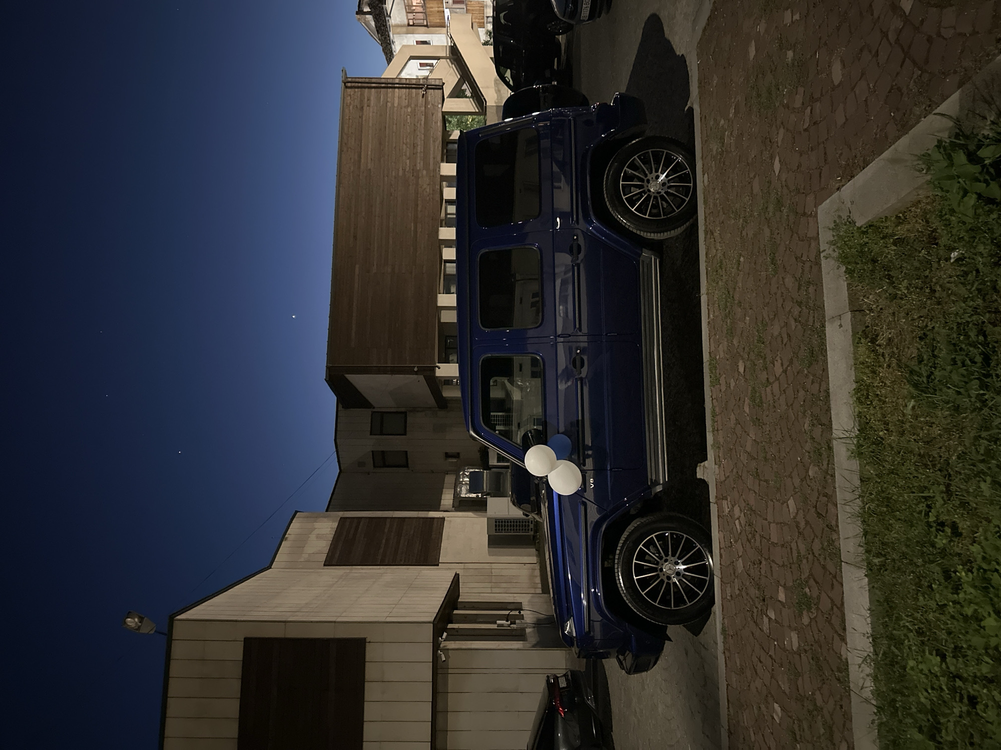 A dark blue Mercedes-Benz G-Class SUV is parked on a cobblestone street, adorned with white and blue balloons attached to the side mirror. The street is bordered by a building constructed of pale brick with dark wooden window treatments, and another building with lighter brick. A street lamp illuminates the corner of the buildings. The time appears to be evening, as indicated by the dark blue sky. The location is in Lovech, Bulgaria.