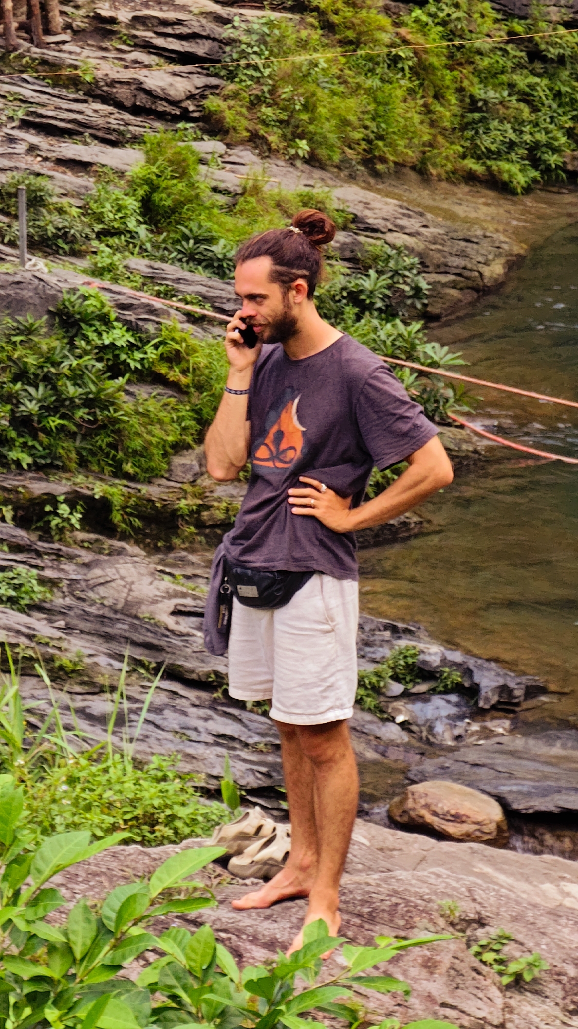 A man stands barefoot on a rock in Du Già Commune, Vietnam, talking on a cell phone. He has a man bun and is wearing a t-shirt with a graphic, white shorts, and a black fanny pack. His shoes are next to him. He is near a body of water with a rope strung across it. The background includes rocky terrain and lush greenery.