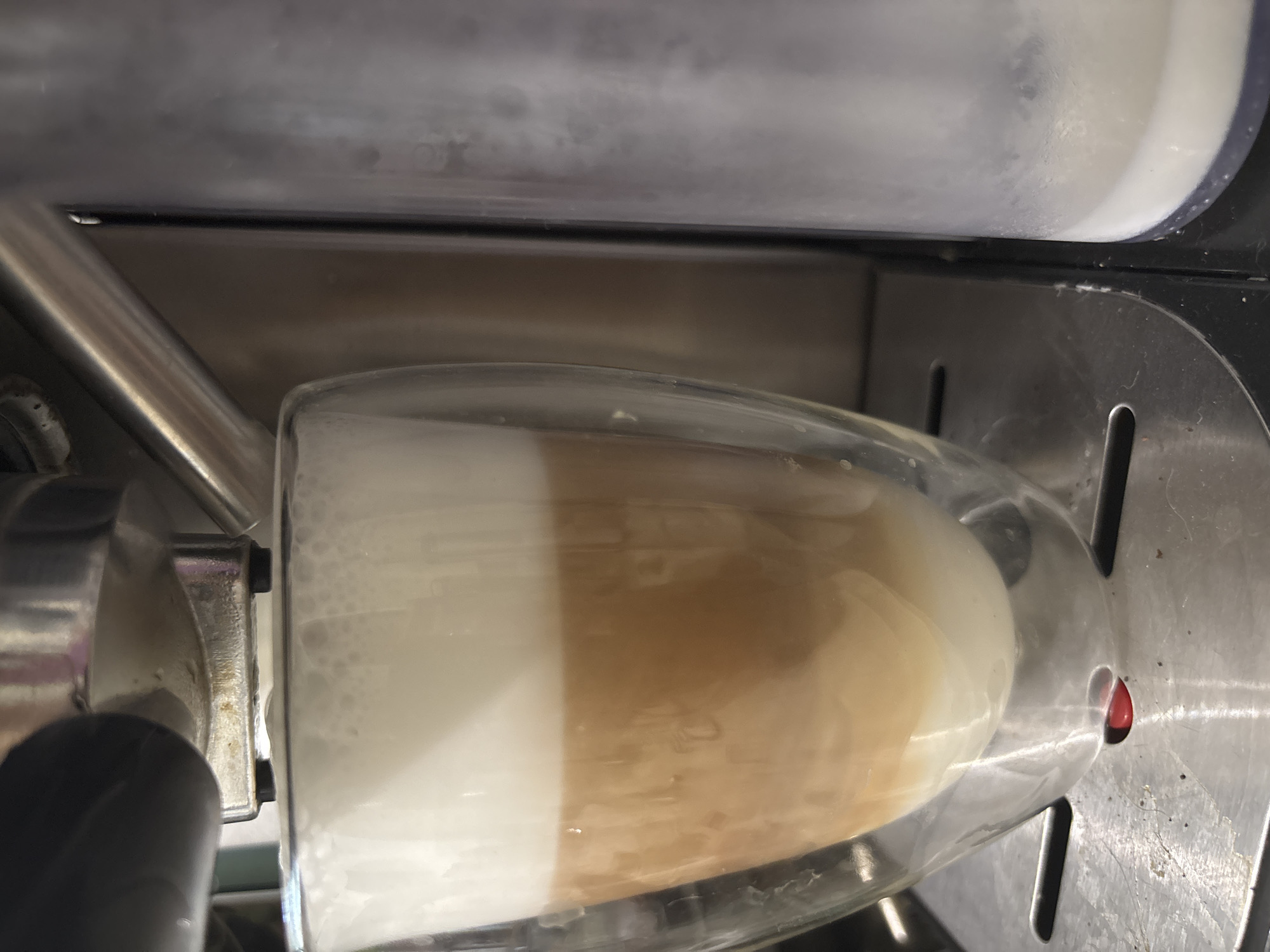 A close-up, eye-level shot captures a glass of a layered coffee drink being prepared by an espresso machine. The drink features distinct layers of white foam at the top, followed by a tan coffee layer, and a milky white base. The glass is transparent and appears to have a double-walled design. The espresso machine's metallic components, including a black handle and a chrome spout, are visible at the top of the frame. To the right, a clear plastic container, possibly for milk or water, is partially visible, showing a white liquid inside. The machine rests on a metallic surface with visible textured details. A small red button or indicator is positioned below the glass. The overall environment appears to be a kitchen or coffee-making area. There is no indication of the location being in Zubov, Russia.