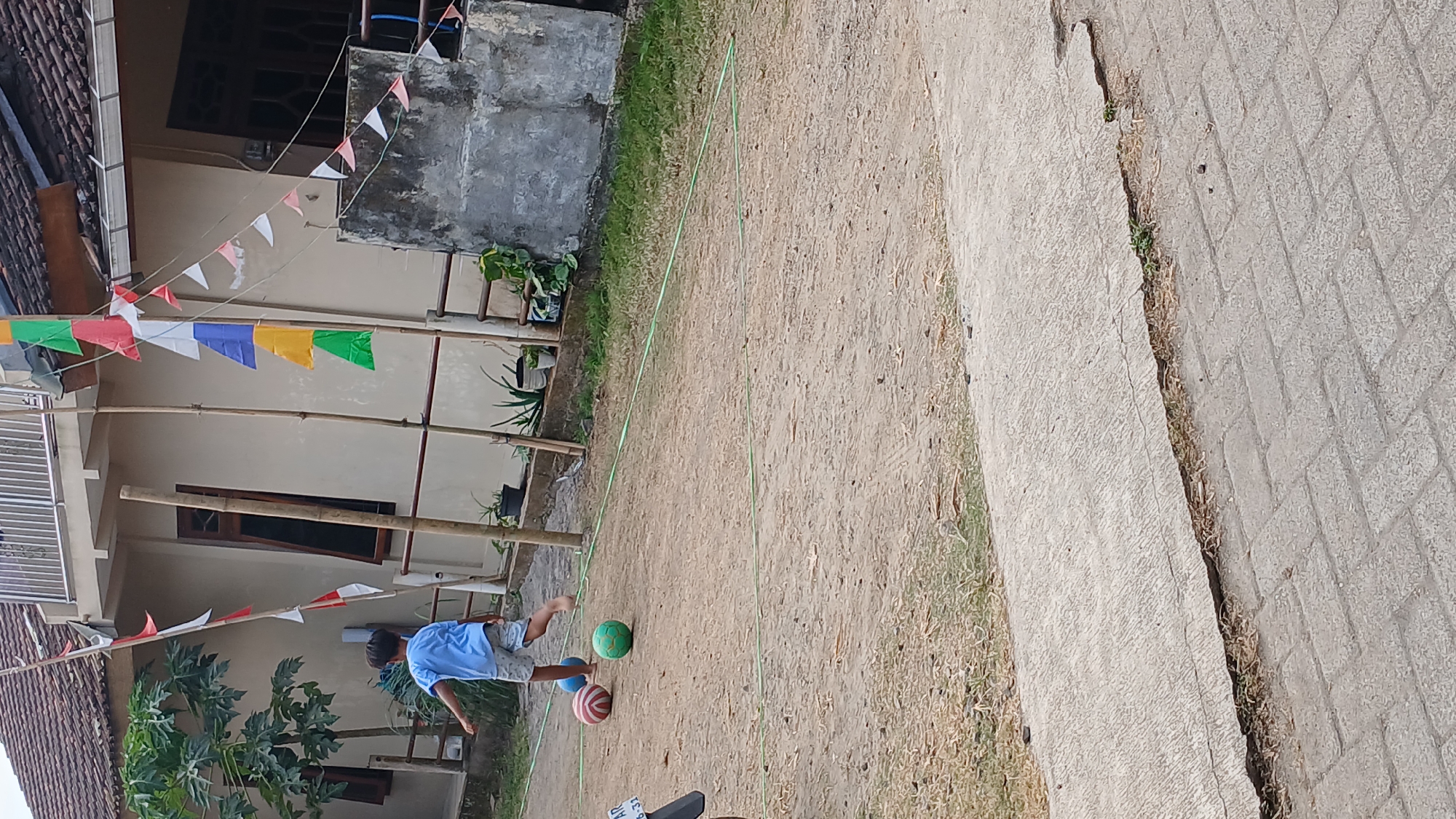 In the city of Mendut, Indonesia, a young boy in a blue t-shirt and grey shorts is playing with two balls on a dirt surface. One ball is green, and the other is red and white striped. The boy is barefoot and appears to be kicking or running towards the balls. The background features a house with a tiled roof and a balcony decorated with colorful triangular flags. Several bamboo poles are also visible, some supporting the flags and others standing independently. Green grass and potted plants are present around the base of the house. A broken concrete pathway is in the foreground.