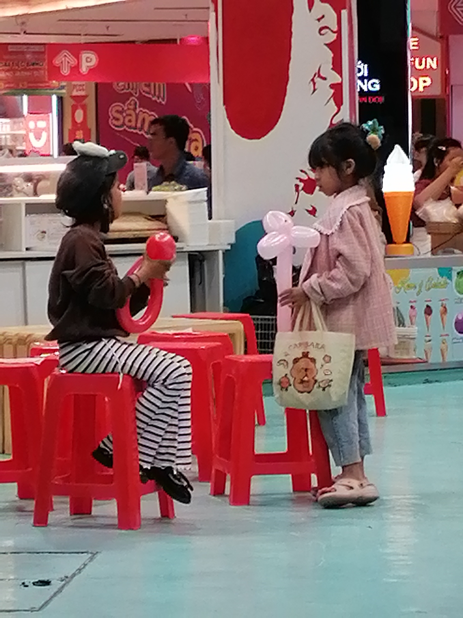 In Da Lat, Vietnam, two young girls are seen sitting on red plastic stools in an indoor setting, possibly a mall or entertainment venue. The girl on the left, wearing a dark long-sleeved shirt and black and white striped pants, is holding a red balloon animal. Her head is adorned with a black hat featuring a white cartoon character. The girl on the right, dressed in a pink jacket and blue jeans, carries a beige tote bag with a cartoon bear design. She is holding a pink balloon animal in her left hand. The background includes a wall with vibrant graphics, a display of ice cream cones, and signs with Vietnamese text. The floor is a light blue-green surface.