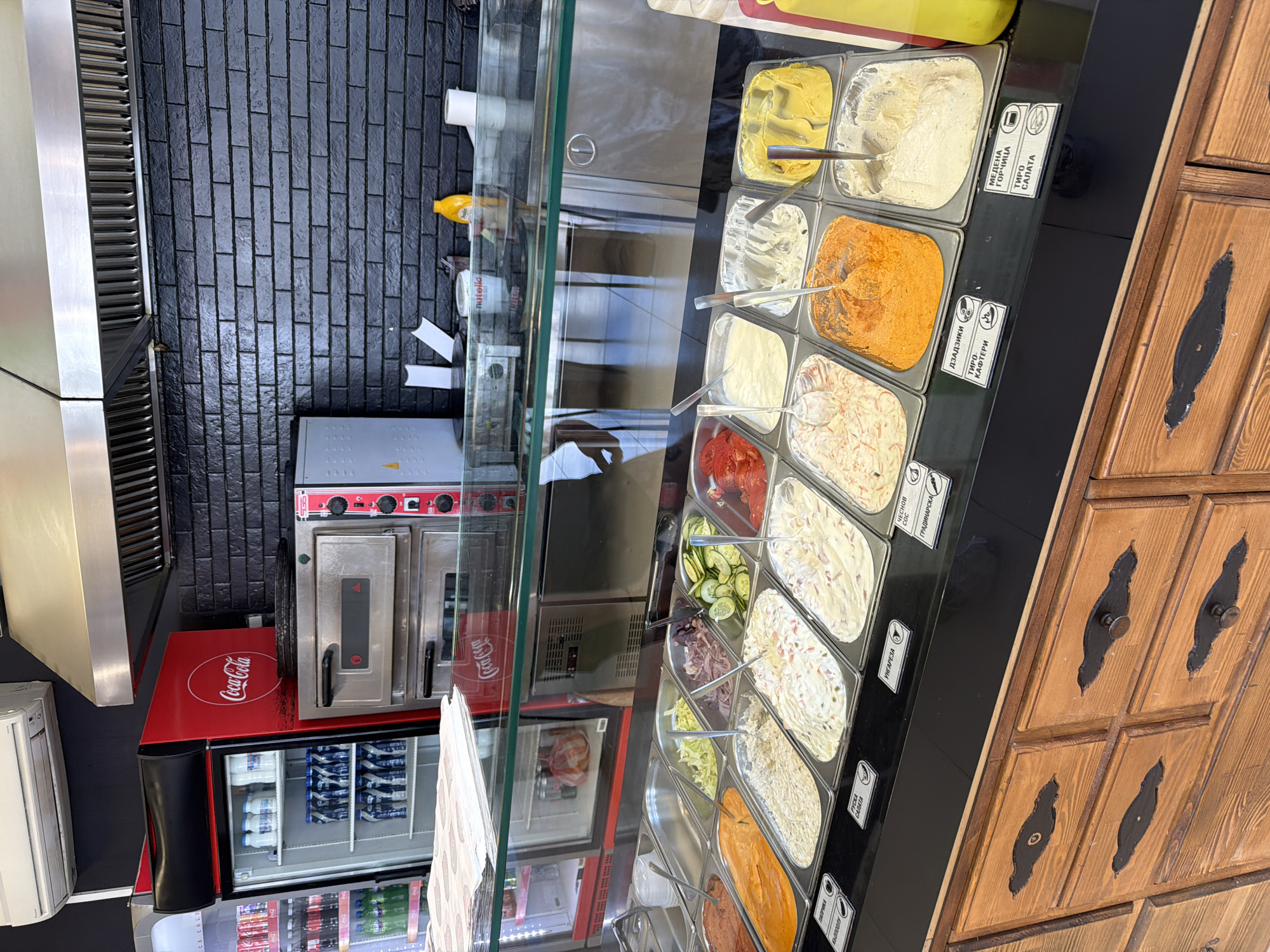 The image presents a view inside a food establishment, likely a fast-food restaurant in Plovdiv, Bulgaria, given the Cyrillic signage. It shows a display case featuring a variety of prepared salads and dips arranged in rectangular containers. Small signs with Cyrillic text, some of which can be translated as "Mustard" or "Russian Salad," identify the contents of each container. Metal serving spoons are placed in each container. A black brick wall is visible in the background, along with a pizza oven and a Coca-Cola branded refrigerator. A wooden cabinet is to the right of the display. The case appears to be situated in a commercial area.