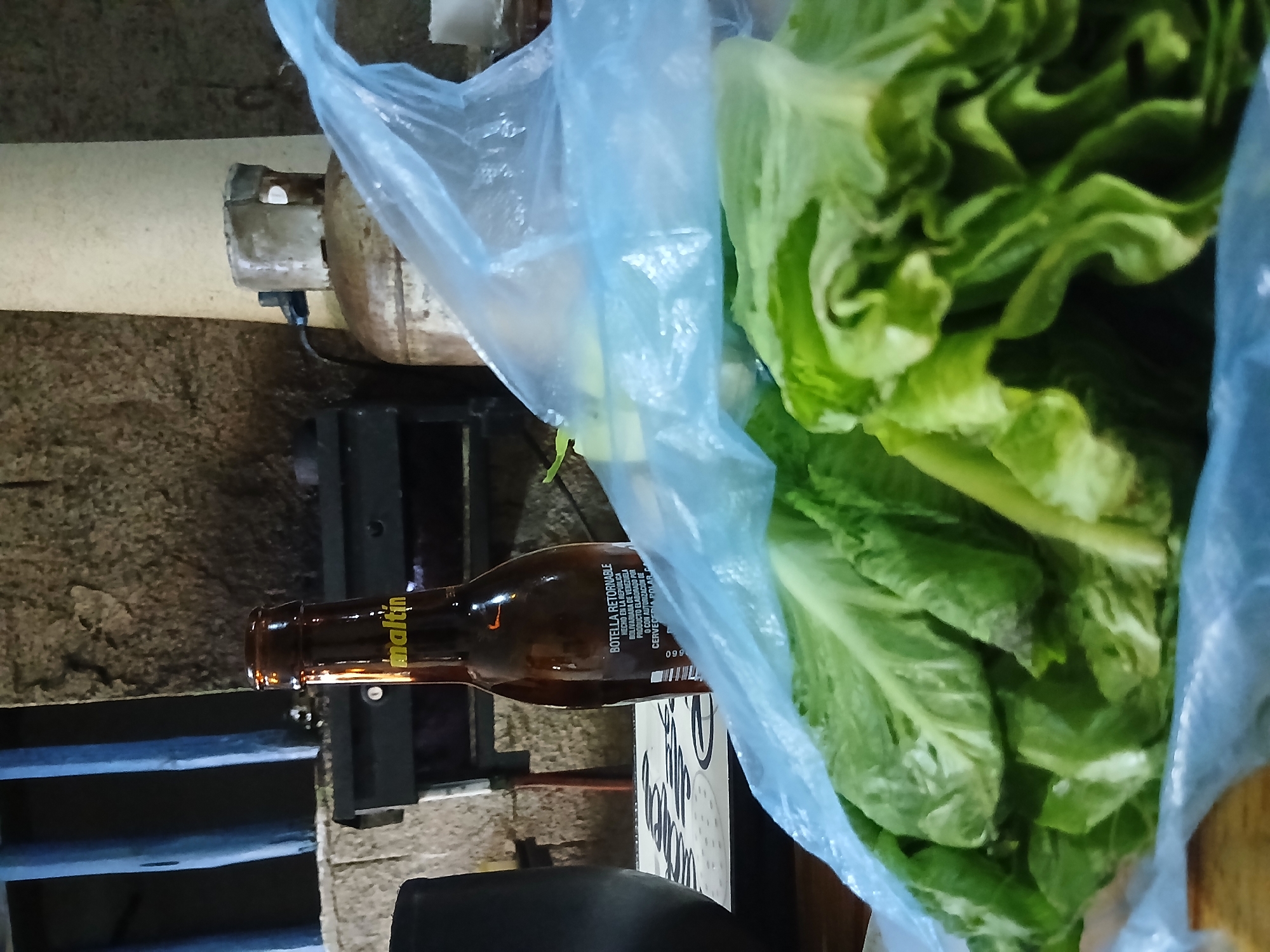 A still life photograph shows a blue plastic grocery bag filled with romaine lettuce next to a brown "Maltin" beer bottle, which rests on a box. To the left of the bottle is a weathered metal frame structure and a horizontal gas tank. The setting appears to be in a storage area or open-air market. The text on the beer bottle mentions "BOTELLA RETORNABLE" and "HECHO EN LA REPÚBLICA BOLIVARIANA DE VENEZUELA" indicating it originates from Venezuela, implying the city of Santa Ana, Venezuela.