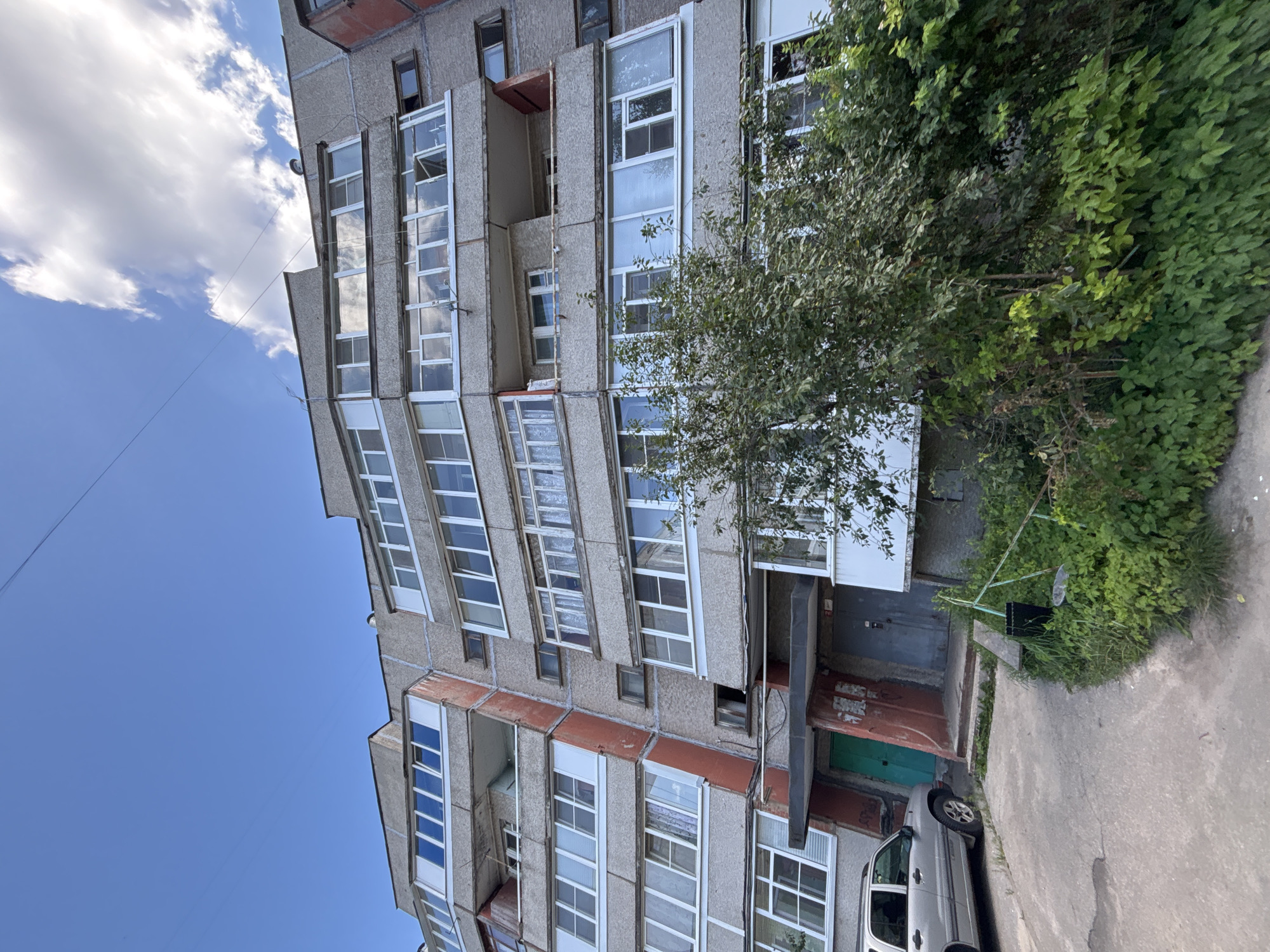 The image depicts a multi-story apartment building in Kstovo, Russia, under a partly cloudy blue sky. The building is constructed of concrete panels and features numerous balconies and windows. A silver SUV is parked on a concrete driveway in front of the building, near two entrances. Dense green foliage surrounds the base of the building, partially obscuring the lower levels. The architecture suggests a Soviet-era apartment block, common in many Russian cities.