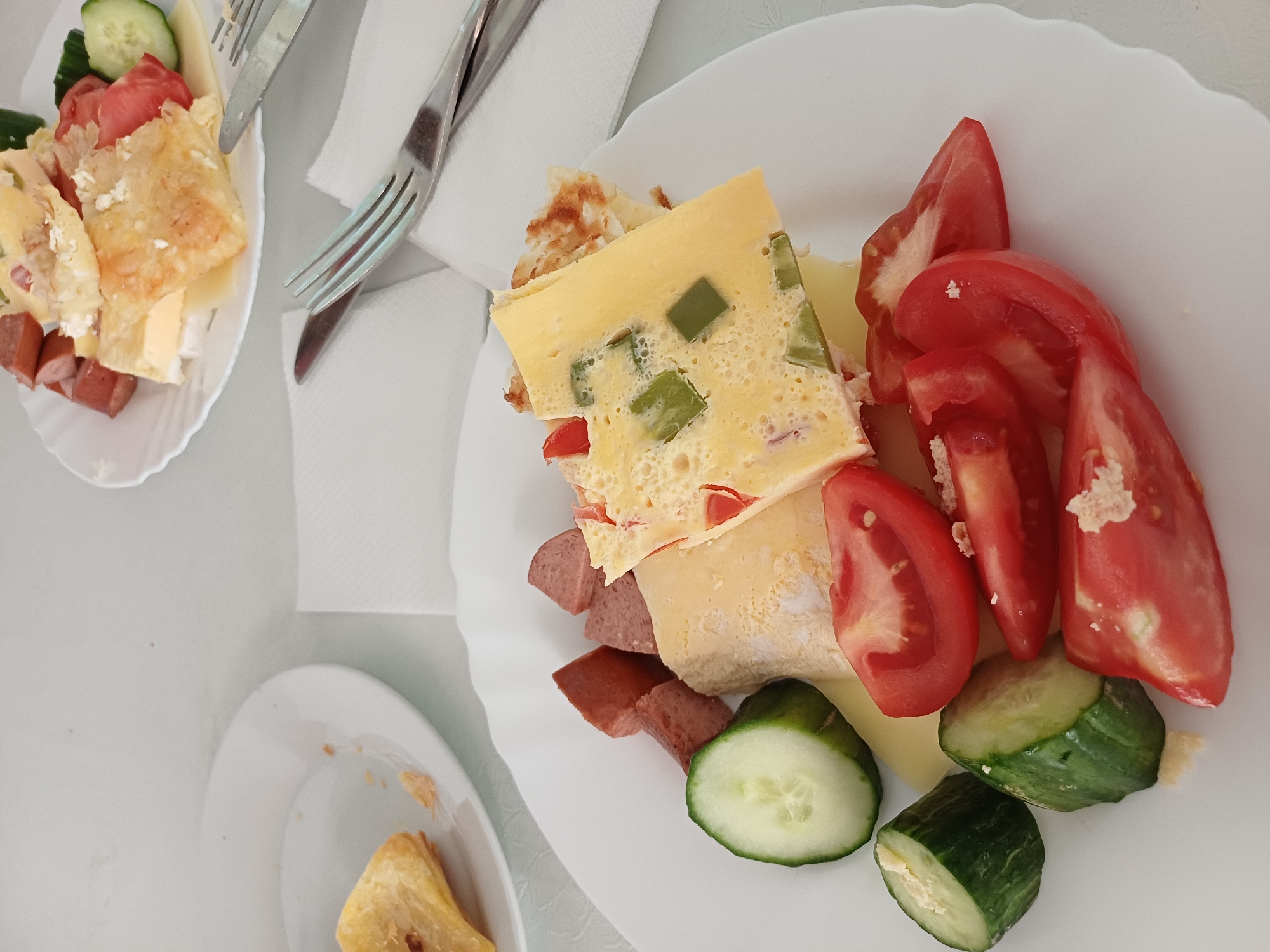 On a white table, two white plates are visible, each containing a breakfast meal. The plate in the foreground displays a slice of omelet with diced green peppers and red tomatoes, accompanied by sliced tomatoes, cucumber rounds, and cut pieces of sausage. Another slice of food, possibly pastry or cheese, is also on the plate. The second plate, slightly out of focus in the background, contains a similar assortment of food, including sausage, cheese, tomato slices, and cucumber. Two forks and a knife are also present on the table, suggesting a meal setting. The overall scene appears to be a breakfast served in Varna, Bulgaria, possibly at a hotel or restaurant, given the organized presentation of the food and the presence of tableware.