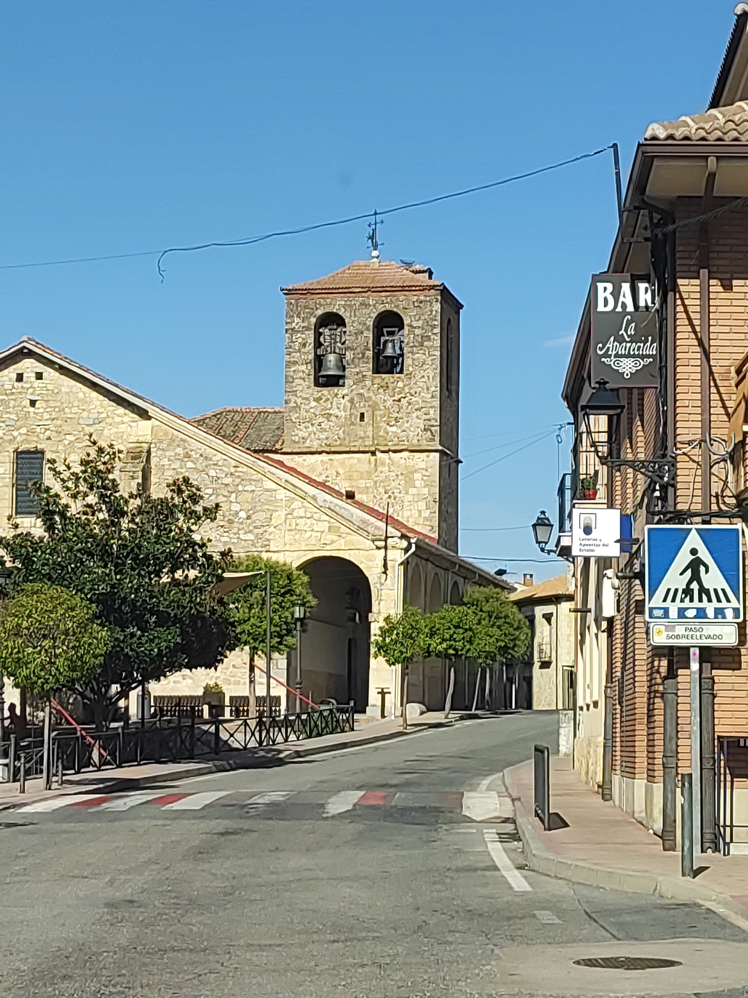 This is a daytime, eye-level, full shot of a street in Valverde Del Majano, Spain, featuring a church with a bell tower and a bar sign. The street is paved and appears to be a two-lane road with a crosswalk marked in white and red stripes. On the left side of the street, a stone church with a tiled roof and an arched entrance is visible, with a large green tree in front of it. The bell tower of the church has two bells. To the right of the street, a brick building has a sign for "BAR La Aperecida." There are also streetlights and a pedestrian crossing sign. The sky is clear and blue. No people are clearly visible.