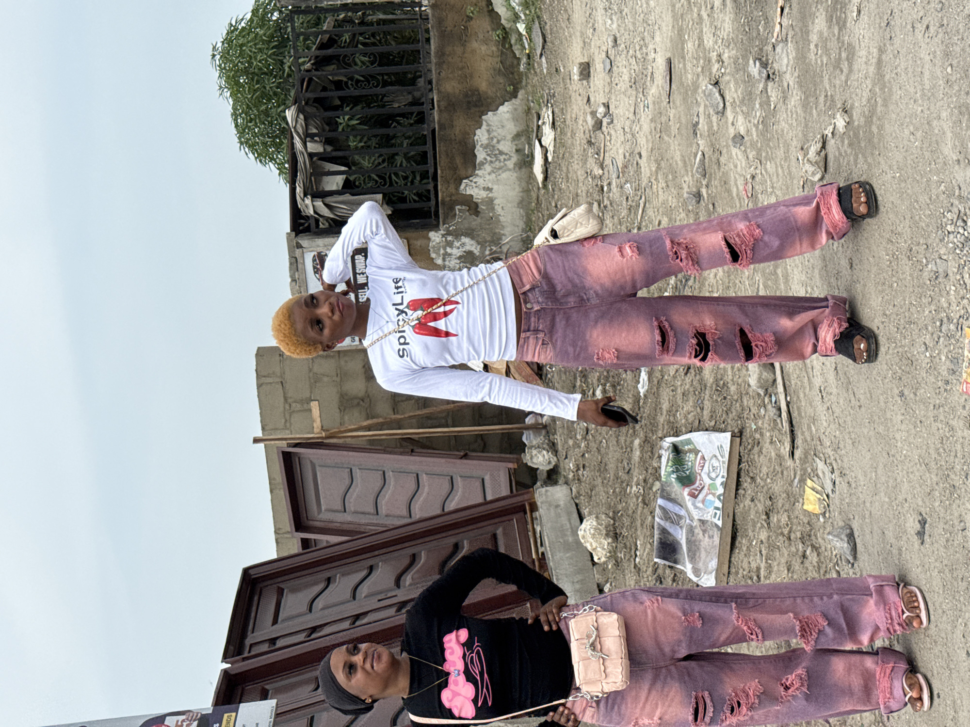 Two women stand on a rough, cracked concrete surface in Ado, Nigeria. One woman wears a white long-sleeved shirt with "Spicy Life" printed on it, while the other is in a black long-sleeved shirt with pink graphics. Both women are wearing matching ripped jeans with pink hues and sandals. A black wrought iron fence and green foliage are visible in the background. Nearby are unpainted wooden doors, construction materials, and scattered refuse.