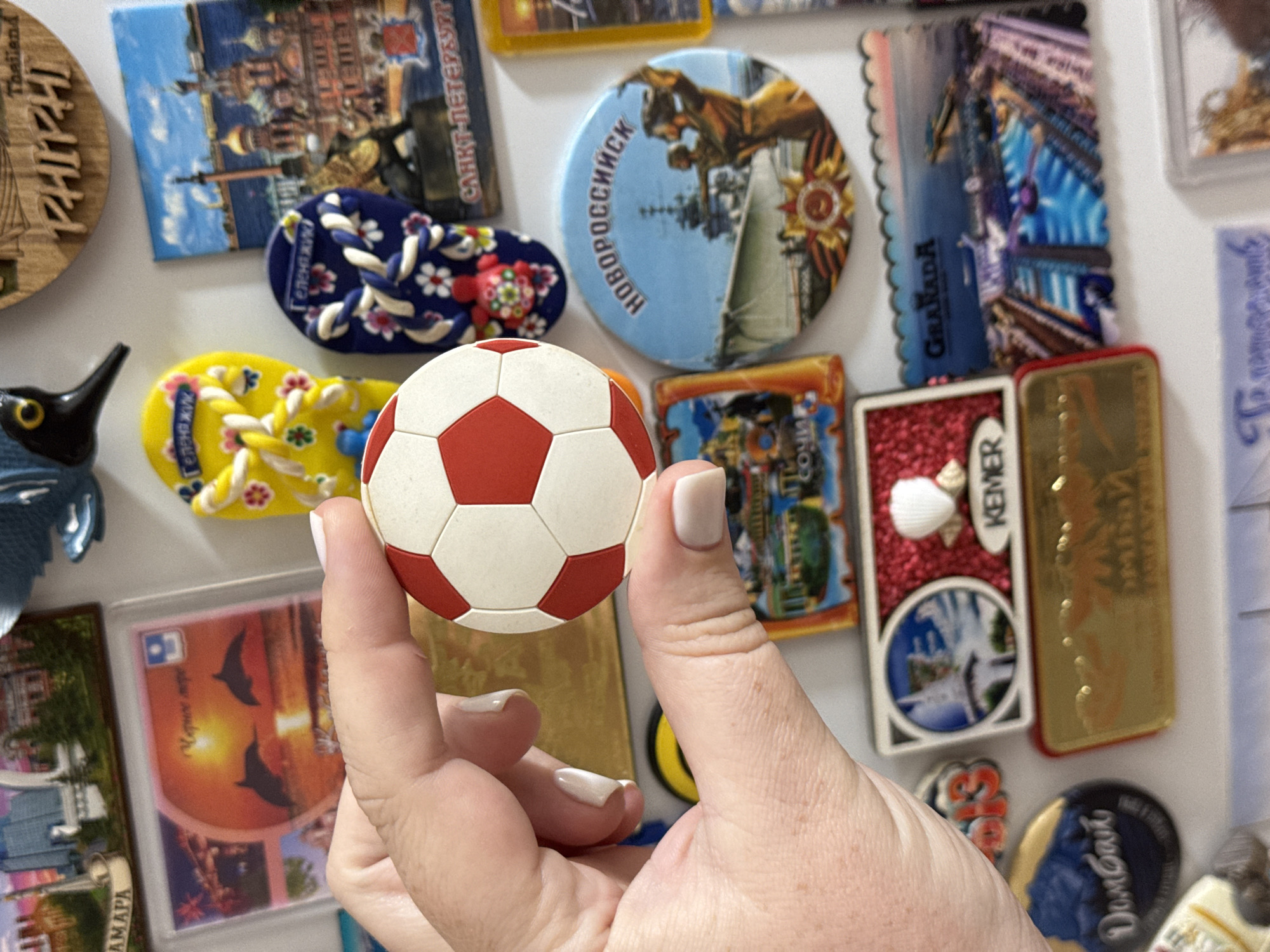 A close-up, eye-level shot shows a hand holding a miniature soccer ball, positioned in front of a collection of colorful refrigerator magnets. The hand, belonging to a person with light-colored fingernails, gently cradles the ball, which is white with red hexagonal panels. The background is a white surface covered with various magnets, some depicting cityscapes, others featuring decorative designs. Notable magnets include a yellow flip-flop with a floral pattern, a blue flip-flop with a similar design, a circular magnet with an image of a ship and figures, and a rectangular magnet with a cityscape and the word "GRANADA". The soccer ball and the hand holding it are the primary focus of the image. The overall scene is indoors, likely on a refrigerator door. There is no visible indication of the city of Mikhaylovsk, Russia, in this image.