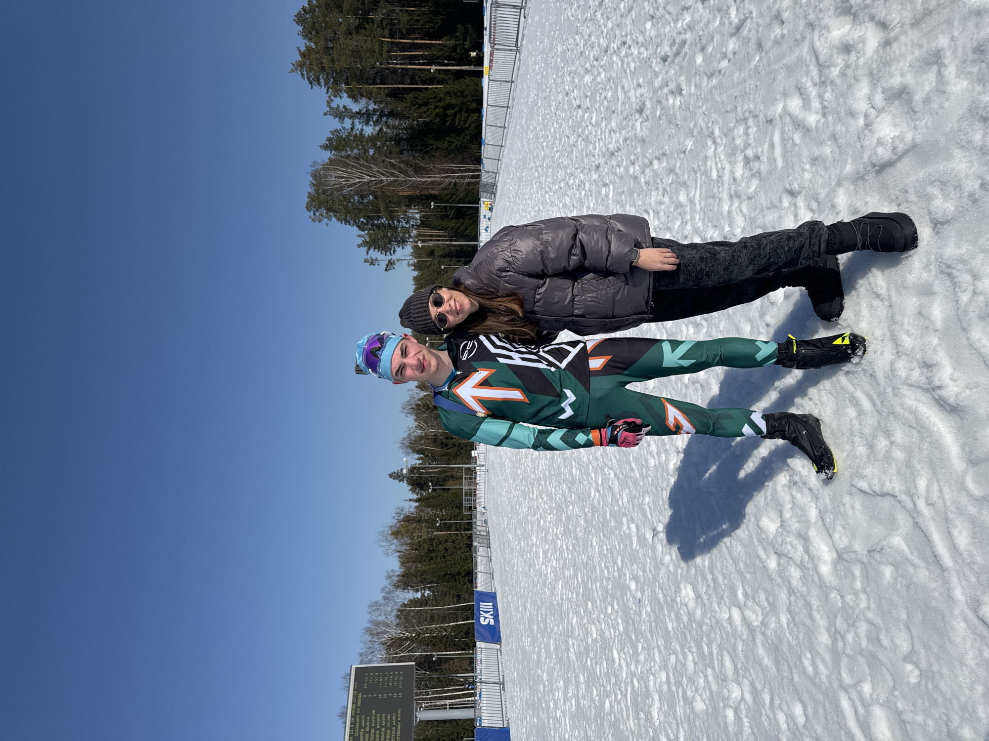 The image depicts two individuals, a male and a female, standing on a snow-covered outdoor course under a clear blue sky in Прикамский, Russia. The male, positioned on the left, wears a multi-colored (green, teal, orange, black, white) athletic ski suit, black ski boots with yellow accents, pink-accented gloves, and a patterned blue and purple beanie. The female, to his right, is dressed in a dark gray puffer jacket, dark patterned pants, black winter boots, a dark gray knit beanie, and sunglasses. Both individuals are smiling towards the camera. The female has her left arm placed behind the male's back. In the background, to the left, a large electronic scoreboard displays race data in Cyrillic, with "SKIII" branding visible on a barrier below. White metal barriers line a snow-covered track extending into the distance. Evergreen and deciduous trees form a treeline behind the course. Bright sunlight illuminates the scene, casting shadows of the individuals on the snow.