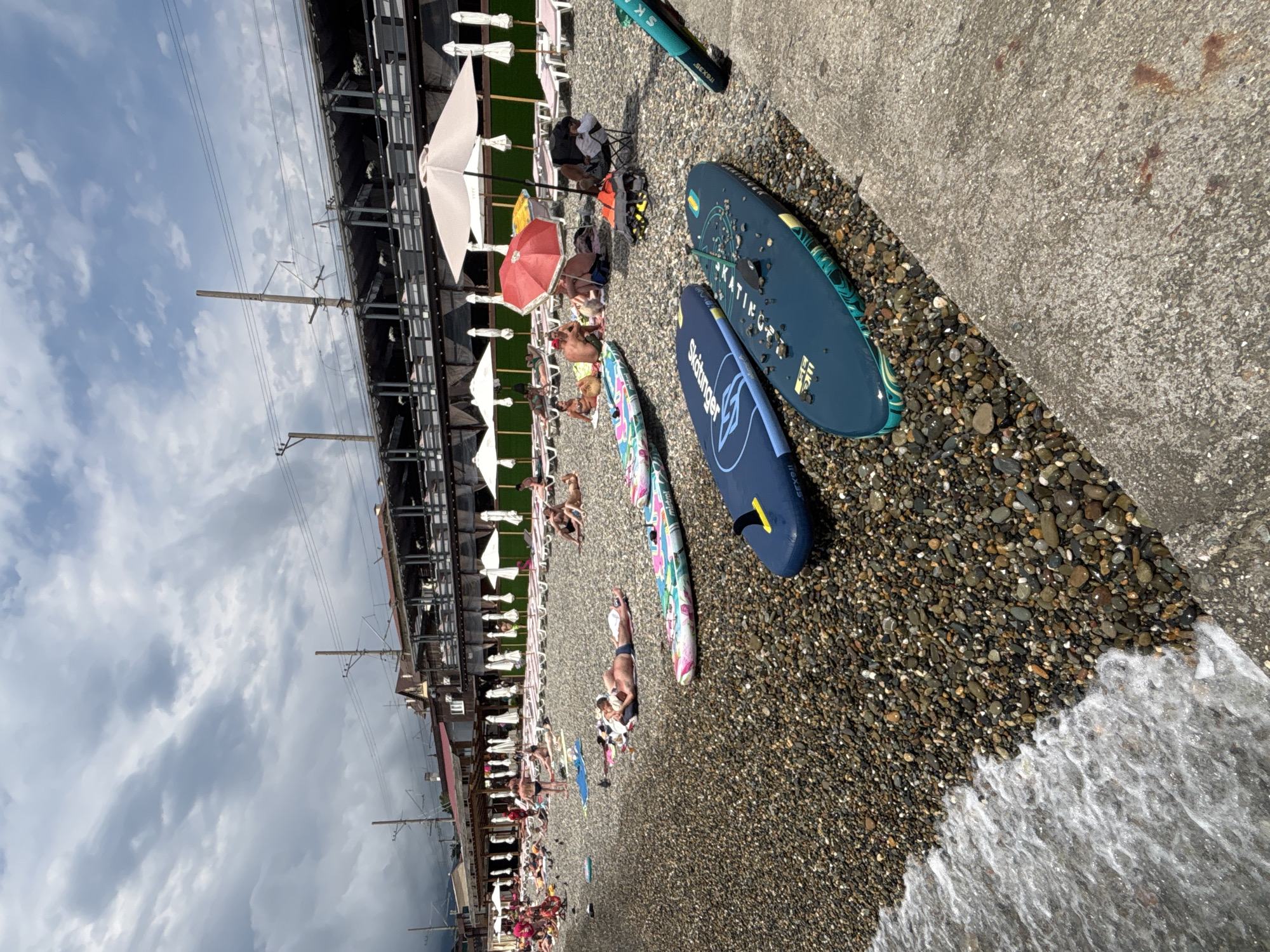 The image shows a pebbled beach in Sochi, Russia, with people relaxing on lounge chairs under umbrellas. Several paddleboards are laid out on the shore, with two in the foreground. The water is visible on the left side of the image. In the background, there is a building with a dark roof and what appears to be a railway track overhead. The sky is cloudy.