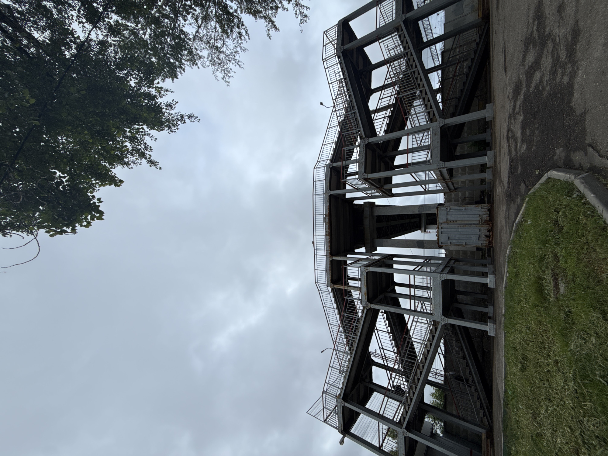 An upward-angled view depicts a multi-level steel structure with open frameworks, staircases, and railings, contrasting with a cloudy sky and some tree foliage on the left. A narrow strip of asphalt and grass borders the structure on the right. The location is Moscow, Russia.