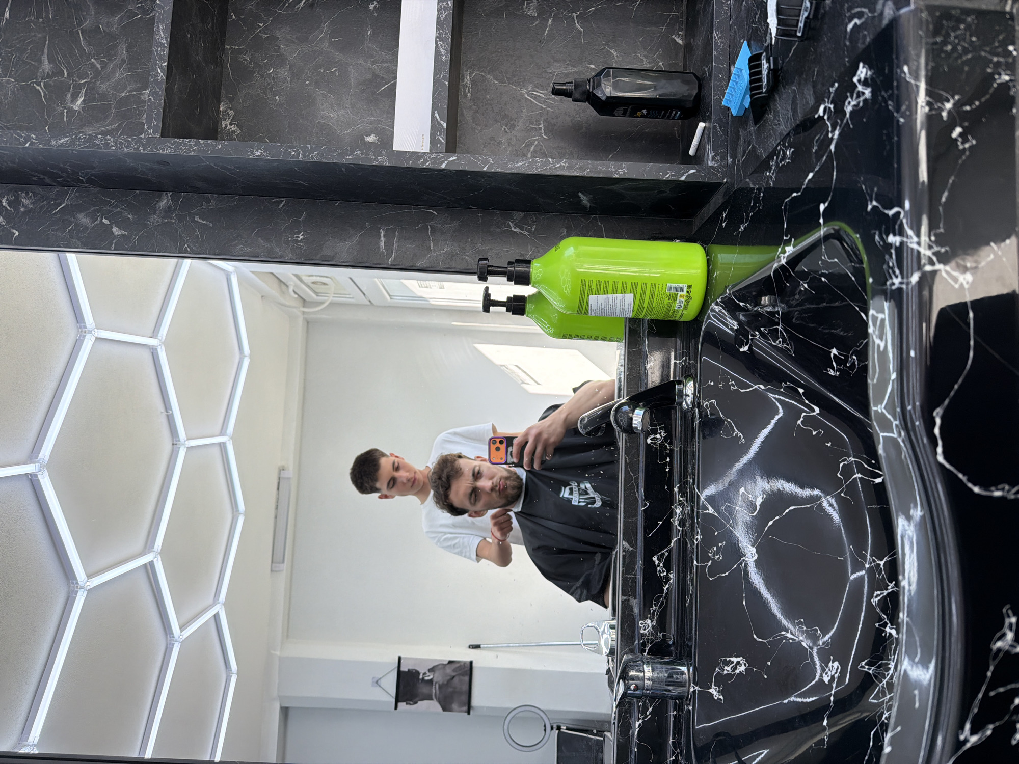 The image displays an interior scene from a barbershop located in Tsarevo, Bulgaria. The central feature is a large rectangular mirror reflecting a male client and his barber, along with parts of the room.
In the reflection, a male client, appearing in his late 20s to early 30s, is seated, wearing a black barber's cape over a dark garment. He has a short, dark, curly beard and short, dark hair. He is holding a smartphone with an orange case in his right hand, positioned to capture a mirror selfie, displaying a slightly pursed-lip expression. Standing behind the client is a younger male barber, likely in his late teens or early 20s, with short dark hair, wearing a white short-sleeved t-shirt. His hands are near the client's head, engaged in hair styling or cutting.
In the immediate foreground, a black vanity counter and integrated sink are visible, characterized by prominent white veining, mimicking marble. Two chrome-finished faucets are installed on the vanity. On the left side of the counter, two identical bright neon-green pump bottles are present. On the right, a black spray bottle, a small white cylindrical object, and a blue hair clipper attachment are placed.
The ceiling, reflected in the mirror, is white and features hexagonal light fixtures composed of illuminated white LED strips. A secondary linear light fixture is mounted on the distant white wall, which also displays a framed black and white poster of a man's head. To the right of the main mirror, a vertical wall panel and a recessed shelf are constructed from the same black material with white veining as the vanity.