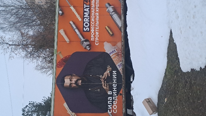 The scene, presented as being in Bel'kovo, Russia, depicts an outdoor environment under an overcast sky. In the foreground, patches of melting snow cover areas of dark, exposed ground, which appears to be dormant grass or dirt. A small, light-brown cardboard box lies horizontally on its side, resting on the ground near a snow patch on the lower left.
Dominating the midground is a large, rectangular advertisement panel, likely mounted on a temporary structure or trailer, positioned above the snow. The advertisement features an orange background. On the left half, a portrait of a man, identifiable as Steven Seagal, is prominently displayed. He wears glasses, a mustache, a goatee, a dark garment resembling a tunic or shirt, and multiple necklaces adorned with pendants. His hands are clasped together at chest level. Below his image, white Cyrillic text reads "СИЛА В СОЕДИНЕНИИ" (Strength in Connection). The right half of the advertisement displays various construction fasteners, including expansion anchors, bolts, and screws. The brand "SORMAT" is visible in white lettering, alongside Cyrillic text "ПРОФЕССИОНАЛЬНЫЙ СТРОИТЕЛЬНЫЙ КРЕПЕЖ" (Professional Construction Fasteners). A partially obscured tube, consistent with a sealant or adhesive product, is also depicted.
The background shows a light, overcast sky. Bare deciduous trees stand on the right, while a dark green coniferous tree is visible on the far left. A light-colored, multi-story building is partially visible through the bare branches on the right side of the image.