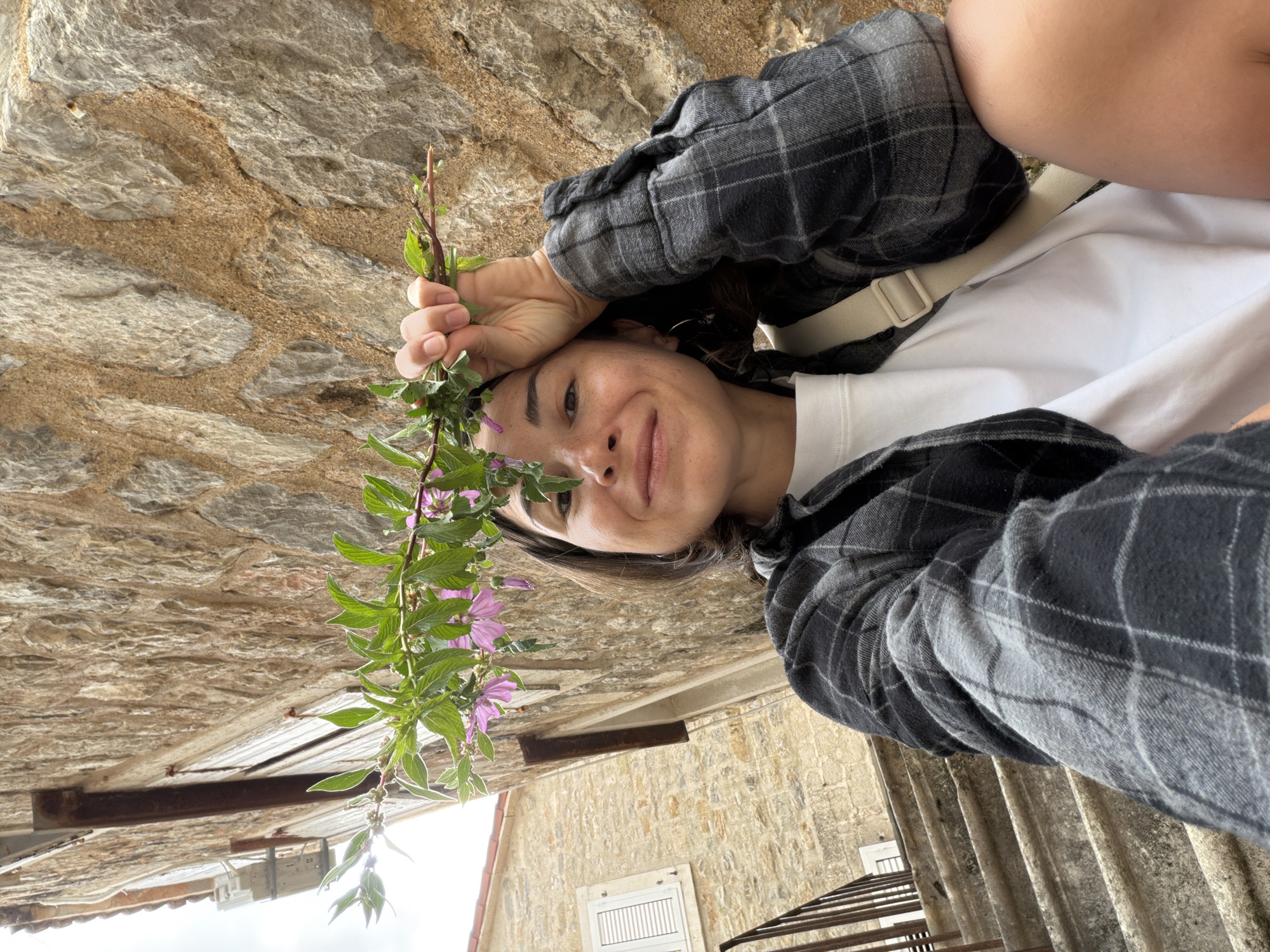 A selfie-style image, rotated, featuring a woman holding a branch with green leaves and pink flowers. She wears a white shirt and a gray and black plaid flannel overshirt, along with a light-colored shoulder strap. The background shows architectural elements: a stone wall, a window with white shutters, metal railings, and a glimpse of the sky. The location is identifiable as Budva, Montenegro based on the architectural style and materials in the background.