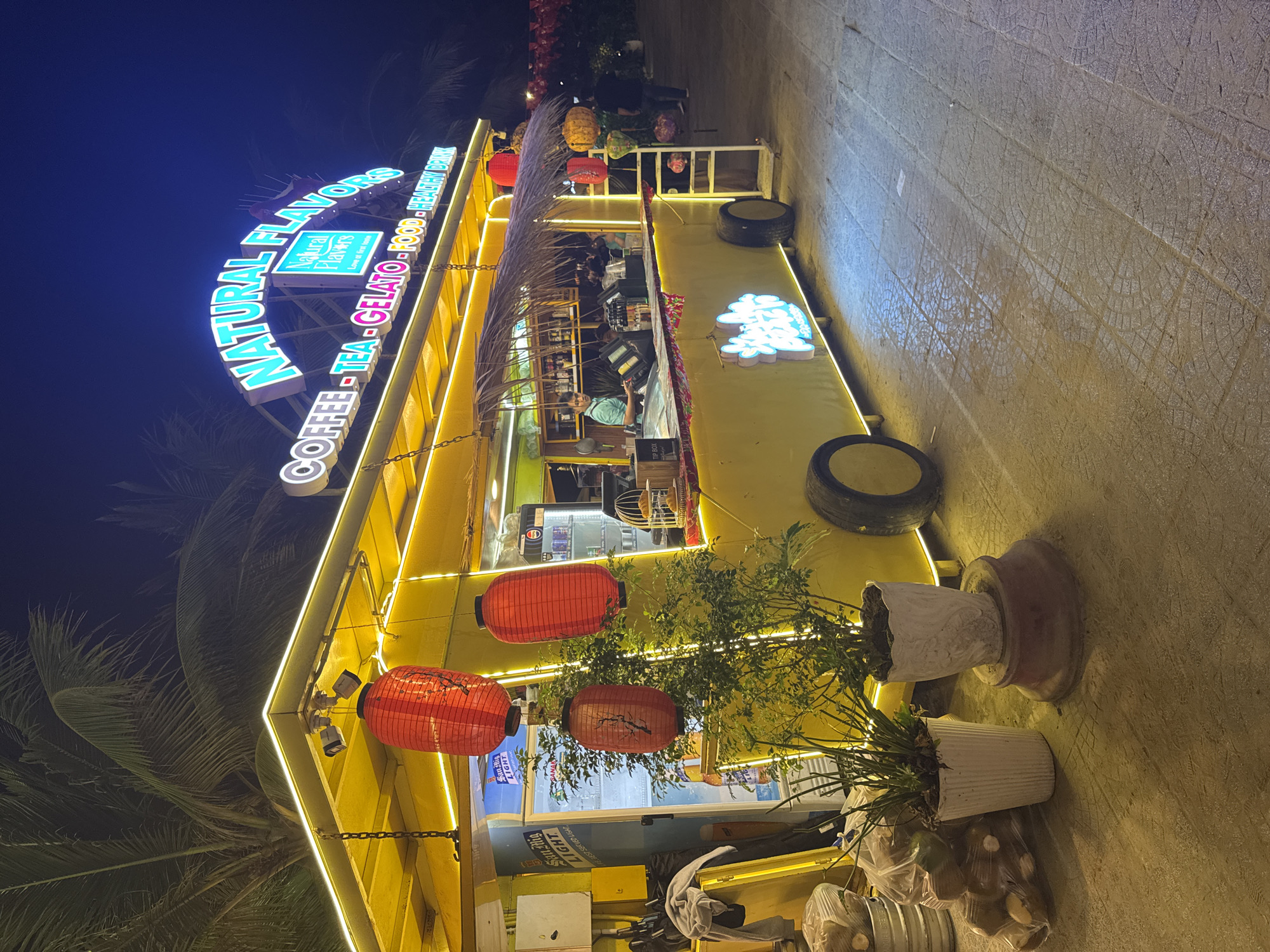 The image depicts a brightly lit "Natural Flavors" coffee, tea, gelato, and food establishment in Đà Nẵng, Vietnam, at night. The structure is yellow with neon trim and features red Chinese lanterns. The sign above reads "Natural Flavors. Coffee - Tea - Gelato - Food - Healthy Drink". There are potted plants and trees decorating the area. Patrons are present inside the establishment. A San Mig Light sign is visible. Tire stacks provide decoration along the side of the structure. The pavement outside is textured.