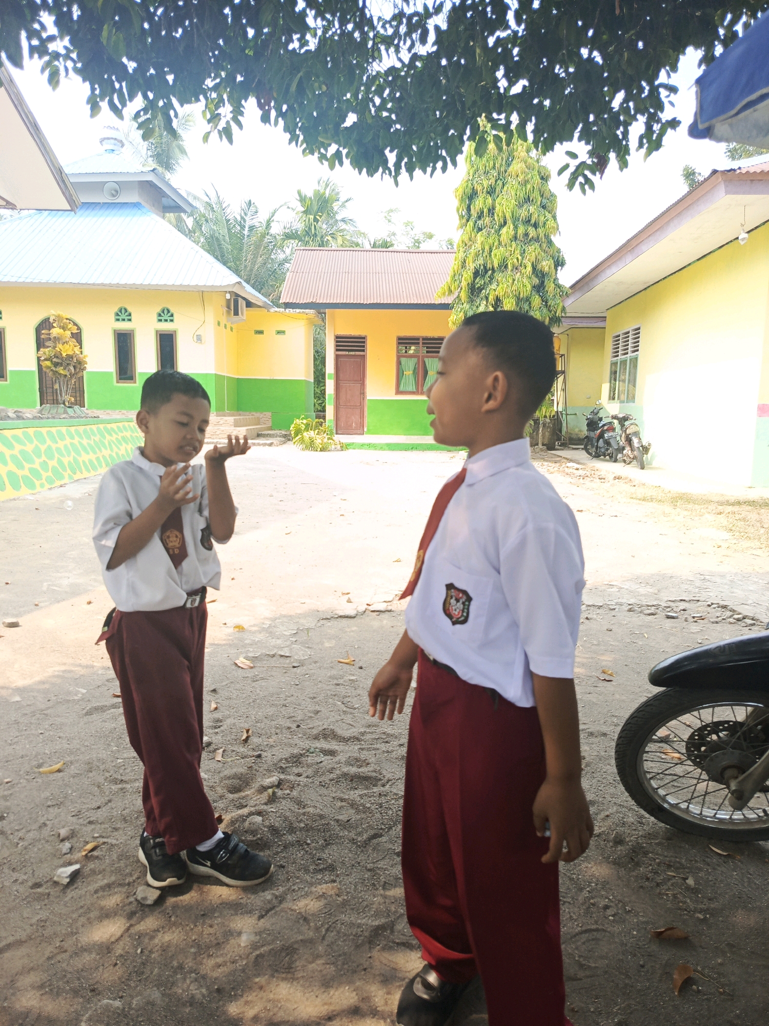 Two young boys in Indonesian school uniforms stand on a dusty ground in front of school buildings. The boy on the left, wearing a white shirt and dark red pants, holds his hands up, appearing to be blowing bubbles or playing. The boy on the right, also in a white shirt and dark red pants, looks to his left, smiling. Behind them, two school buildings are visible. One building is yellow with a light blue roof and green trim. The other building is also yellow with a brown roof. A motorcycle is parked to the right of the image, with its front wheel and part of the handlebars visible. Trees and foliage are present in the background, with some leaves obscuring the top of the image. The scene appears to be set in an outdoor area of a school in the city of Karya Dadi, Indonesia.