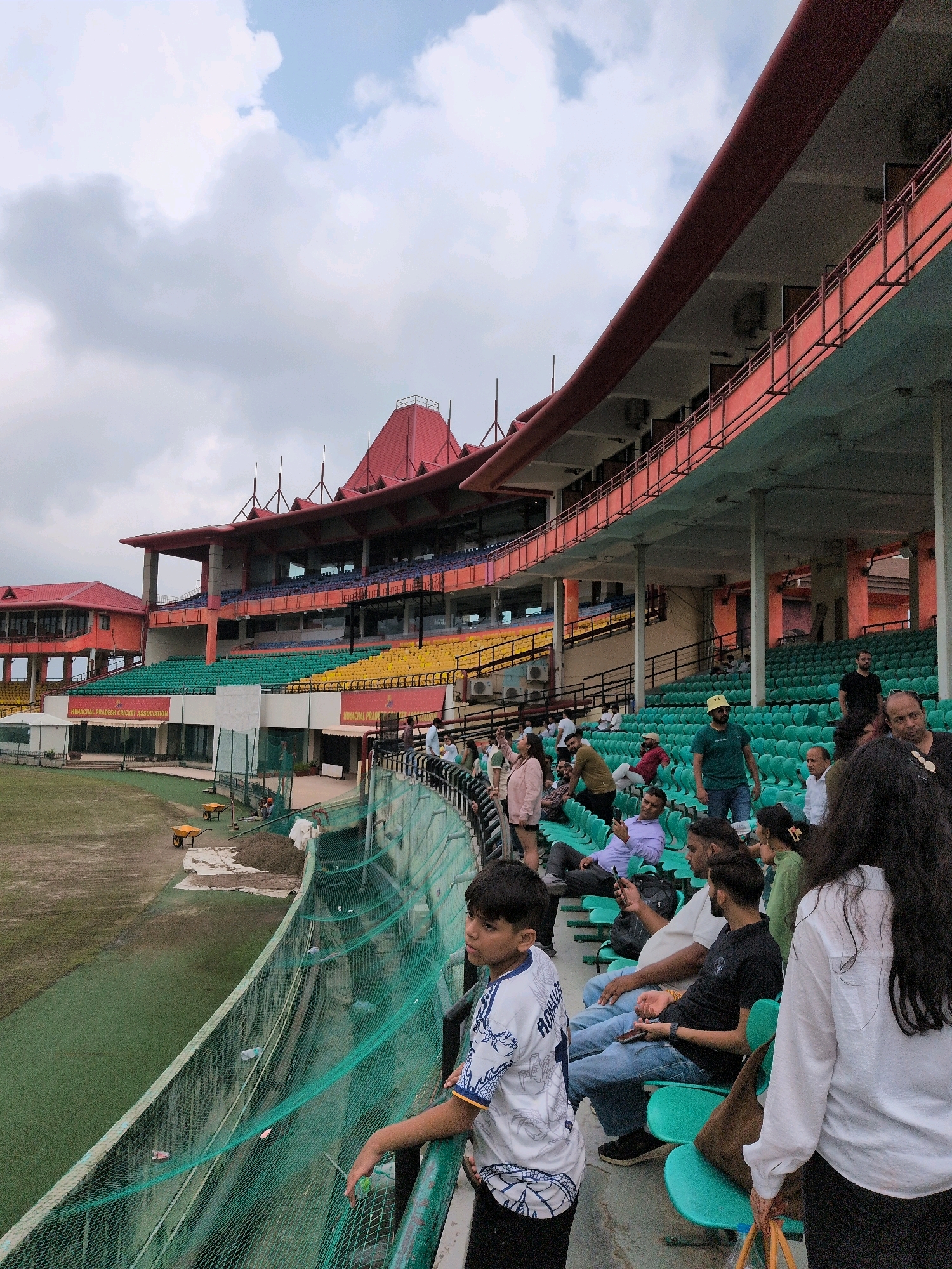 This image captures a scene at the Himachal Pradesh Cricket Association Stadium in Dharamshala, India. The stadium, known for its picturesque setting in the Himalayas, features multi-tiered seating in green, yellow, and blue. The architecture is characterized by red-tiled roofs and prominent red railings.
In the foreground, a young boy wearing a white and blue cricket jersey with the name "RONALDO" visible on the back is looking towards the field. He is standing near a green net barrier. Behind him, several spectators are seated on teal-colored chairs, some looking towards the field and others engaged in conversation or looking at their phones.
Further back, more rows of empty green seats are visible, indicating that some sections of the stadium might not be fully occupied. The field of play, covered in lush green grass, is visible on the left side of the image. The sky is overcast with white clouds. Construction or maintenance activity is suggested by wheelbarrows and piles of soil near the field. The overall atmosphere appears to be one of casual observation, possibly during a break in a sporting event or practice.