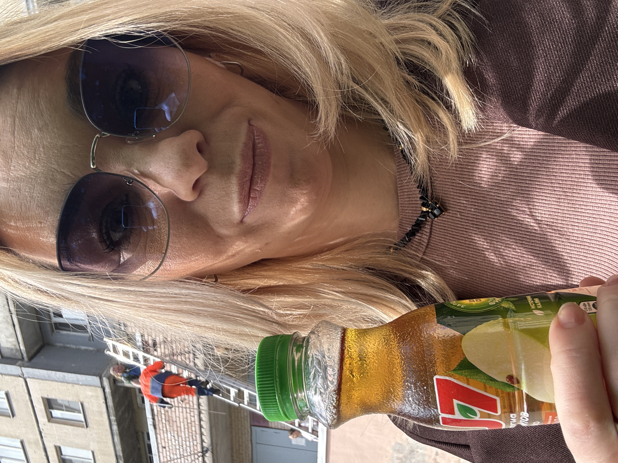 A woman with blonde hair wearing sunglasses is holding a bottle of 7 Up juice. In the background is a building and a man on a ladder. The location is the city of Saint Petersburg, Russia.