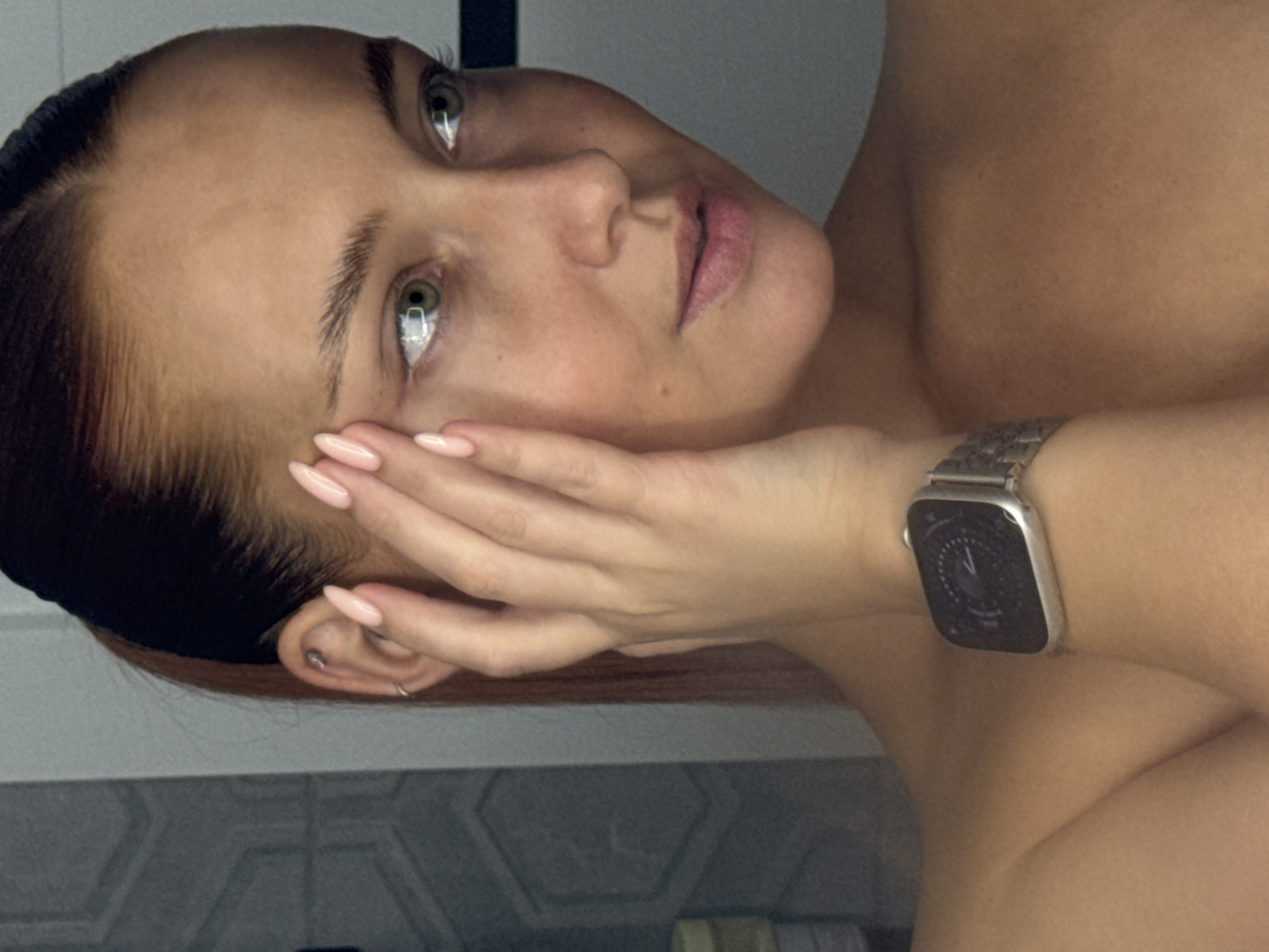 A woman's face is the primary focus of this close-up image. She has light skin, green eyes, and dark hair styled back. Her left hand is raised to her face, with her fingers gently touching her cheek. She is wearing a silver smartwatch on her left wrist. The background is nondescript and consists of grey and white surfaces, suggesting a bathroom or similarly tiled area. The location is unspecified.