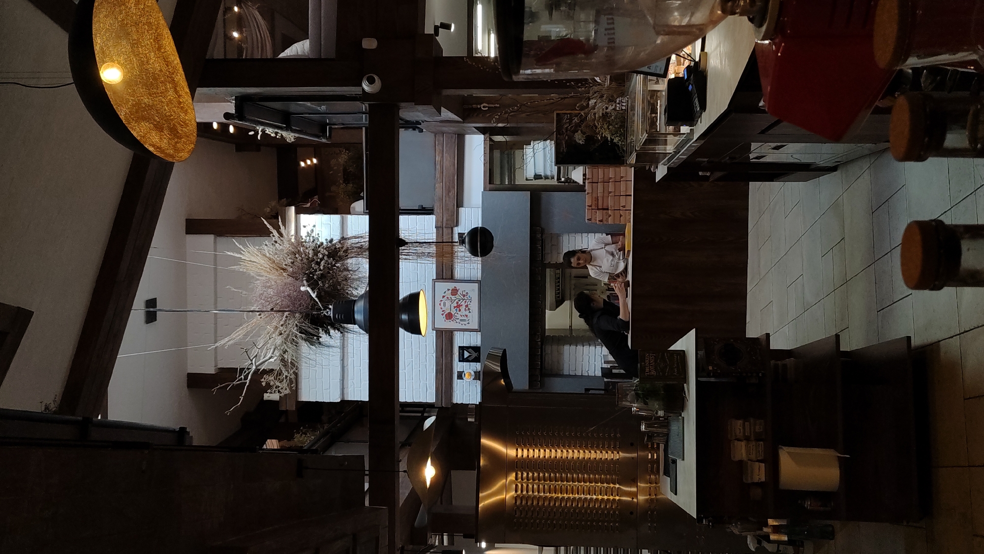 The image depicts the interior of a restaurant or cafe, possibly located in Plovdiv, Bulgaria, given the overall aesthetic. The scene is dimly lit, with a focus on the central area of the establishment. In the background, two women are visible behind a counter. One woman, wearing a black top, appears to be interacting with the other woman who is wearing a white apron, suggesting they are staff members. They are positioned near a brick wall that seems to be part of a decorative fireplace or oven feature.
Dominating the foreground and mid-ground are various elements of the restaurant's decor and operational setup. A large, metallic appliance, possibly a pizza oven or a food preparation station, is visible on the left. In front of it, a multi-tiered shelf unit holds various items, including what appear to be paper products and bottles. A long counter stretches across the scene, with a coffee machine or blender visible on the far right. Several boxes, stacked neatly, are also present on the counter, possibly for takeaway orders.
The ceiling features exposed wooden beams and multiple light fixtures, including a prominent, large, golden lampshade on the right and two black pendant lights in the center. A large dried floral arrangement hangs from the ceiling in the central area, adding a rustic touch to the ambiance. The floor is tiled with large square tiles. The overall impression is one of a cozy, somewhat rustic, and possibly artisanal establishment.